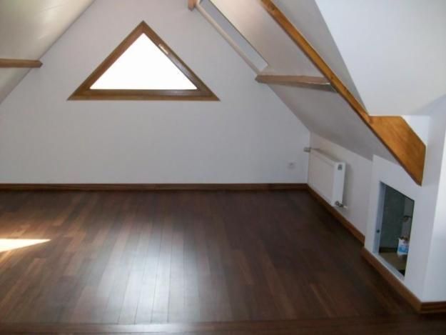 Pièce mansardée avec parquet foncé, fenêtre triangulaire, murs blancs et poutres apparentes.
