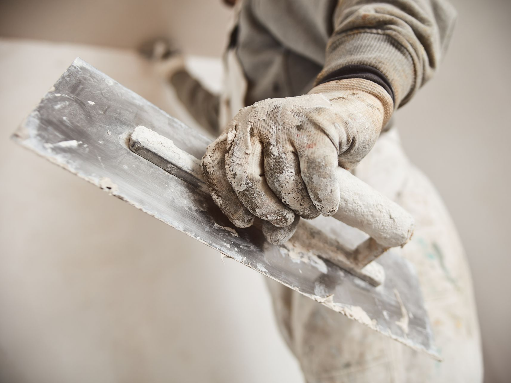 Personne portant des gants de travail et tenant une truelle, appliquant du plâtre sur un mur.