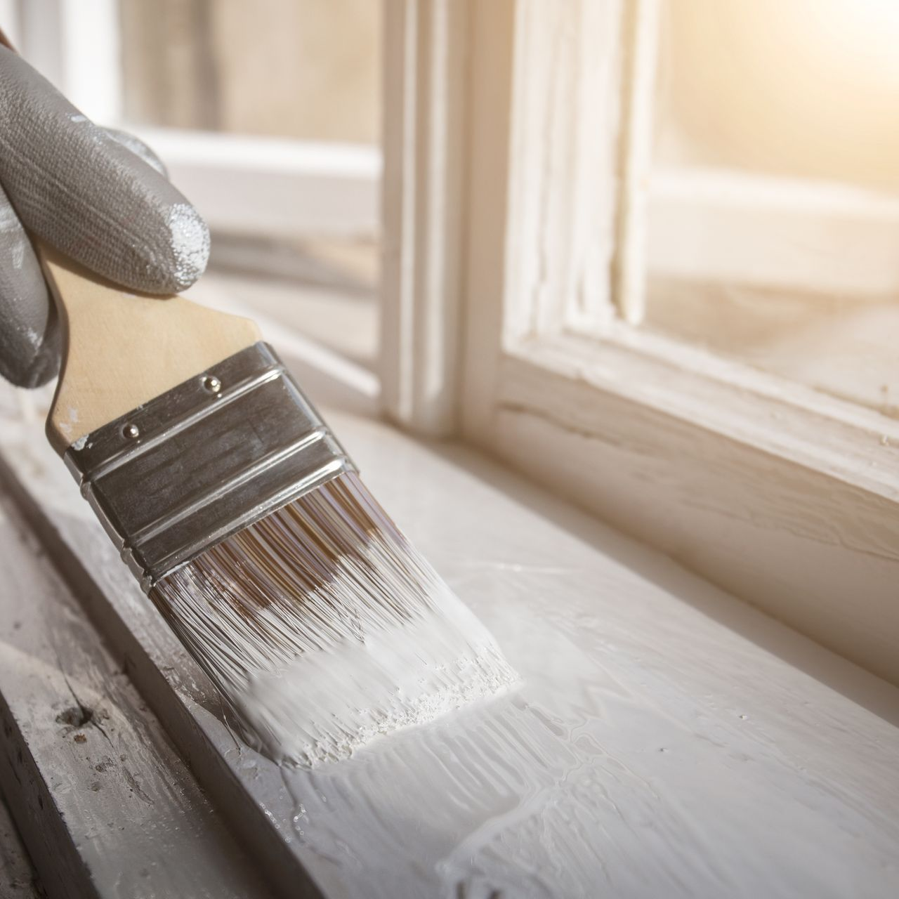 Main gantée peignant un cadre de fenêtre blanc avec un pinceau et de la peinture blanche.