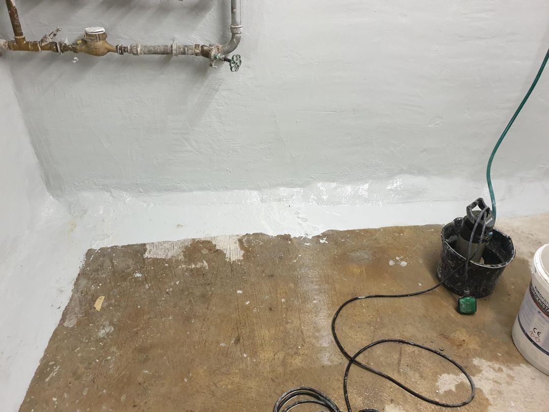 Keller mit weiß gestrichenen Wänden, Betonboden und einer Pumpe mit Schlauch. Wasserleitungen an der Wand.