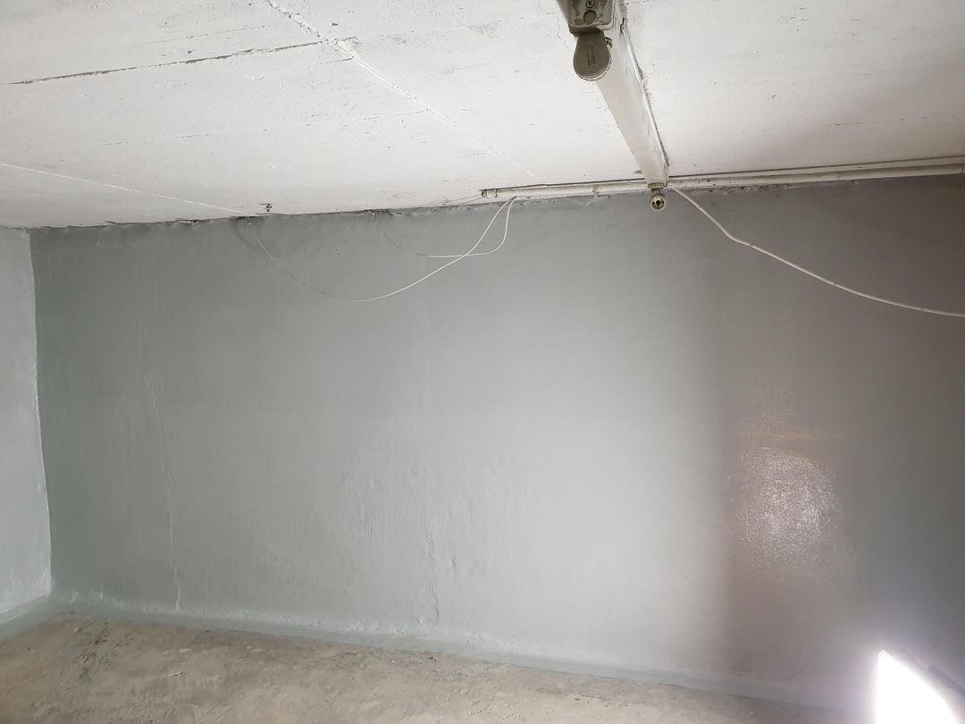 Grau gestrichene Innenwand mit weißer Decke und Betonboden. Einige elektrische Leitungen sind sichtbar.