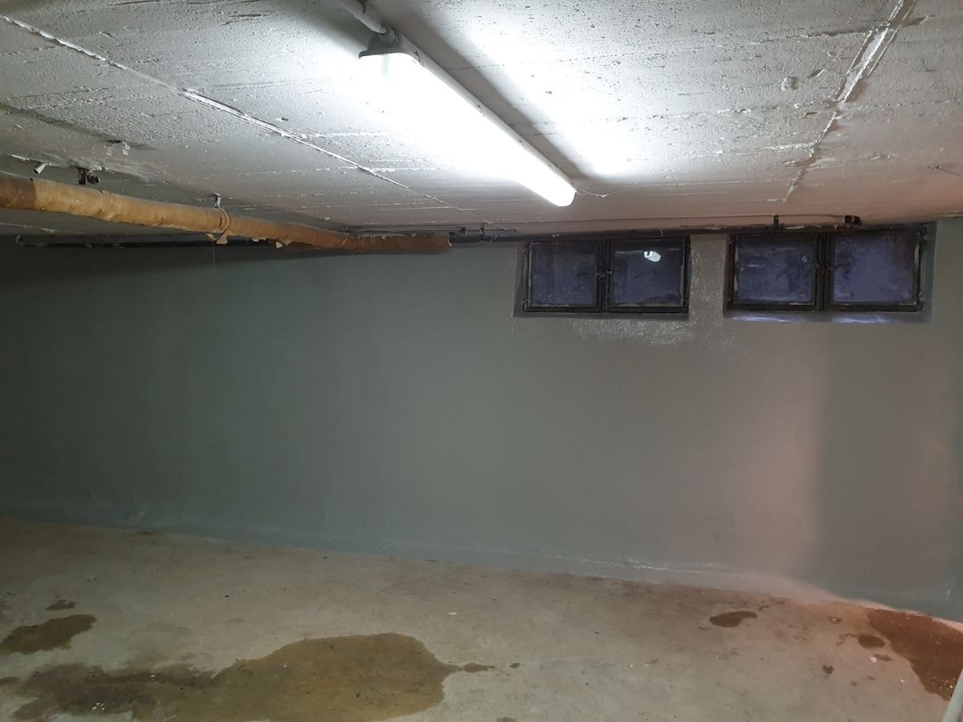 Kellerraum mit grauen Wänden und Betondecke, mit zwei kleinen Fenstern und einem nassen Boden.