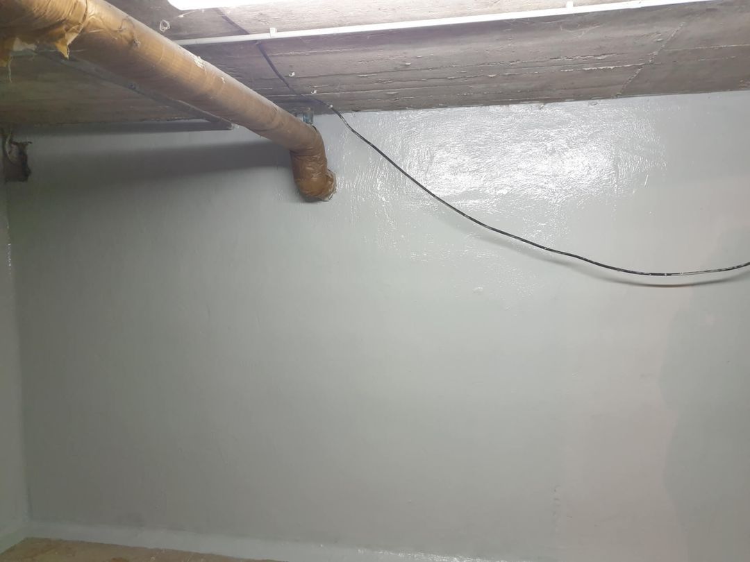 Eine grau gestrichene Kellerwand, an der ein Rohr, Kabel und die Decke sichtbar sind.
