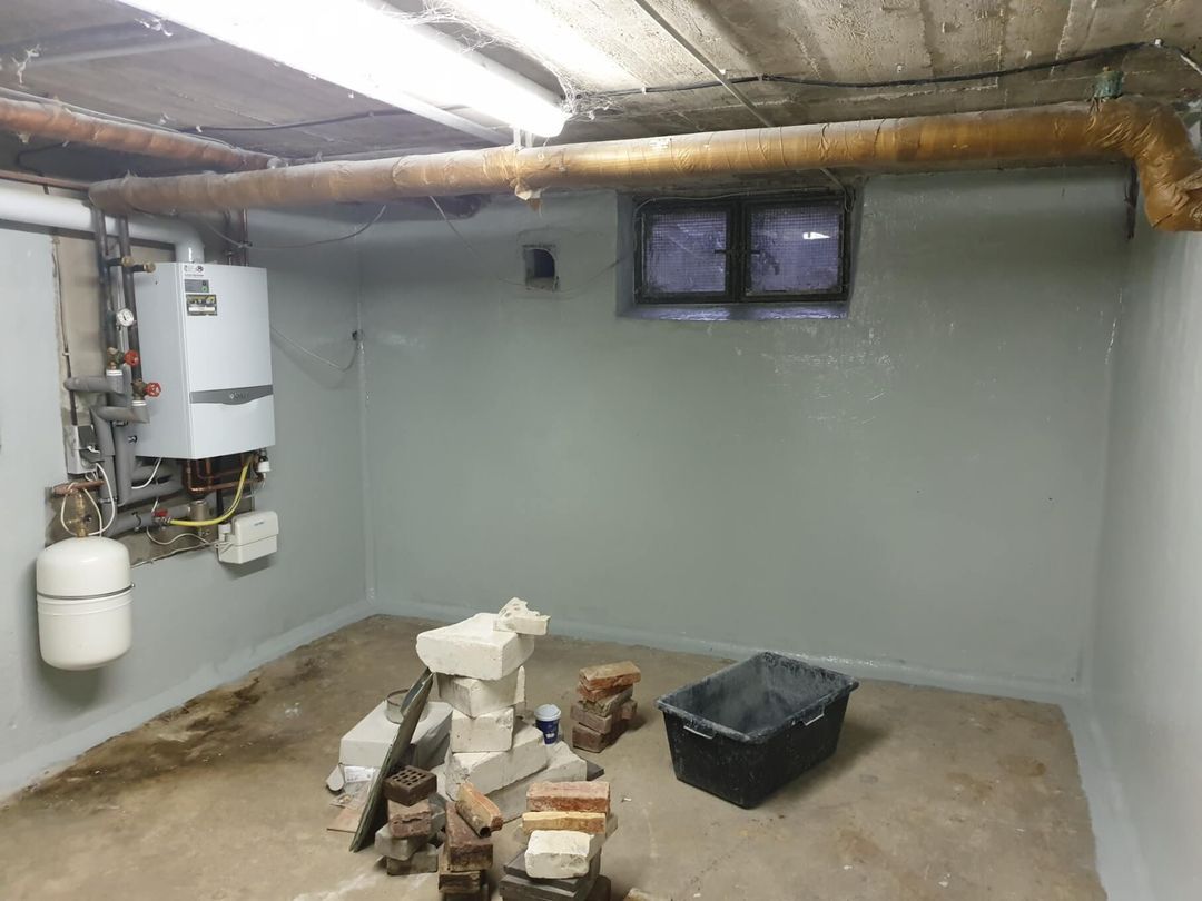 Keller mit Heizkessel, kleinem Fenster, schwarzer Tonne, Ziegeln und Zementboden.