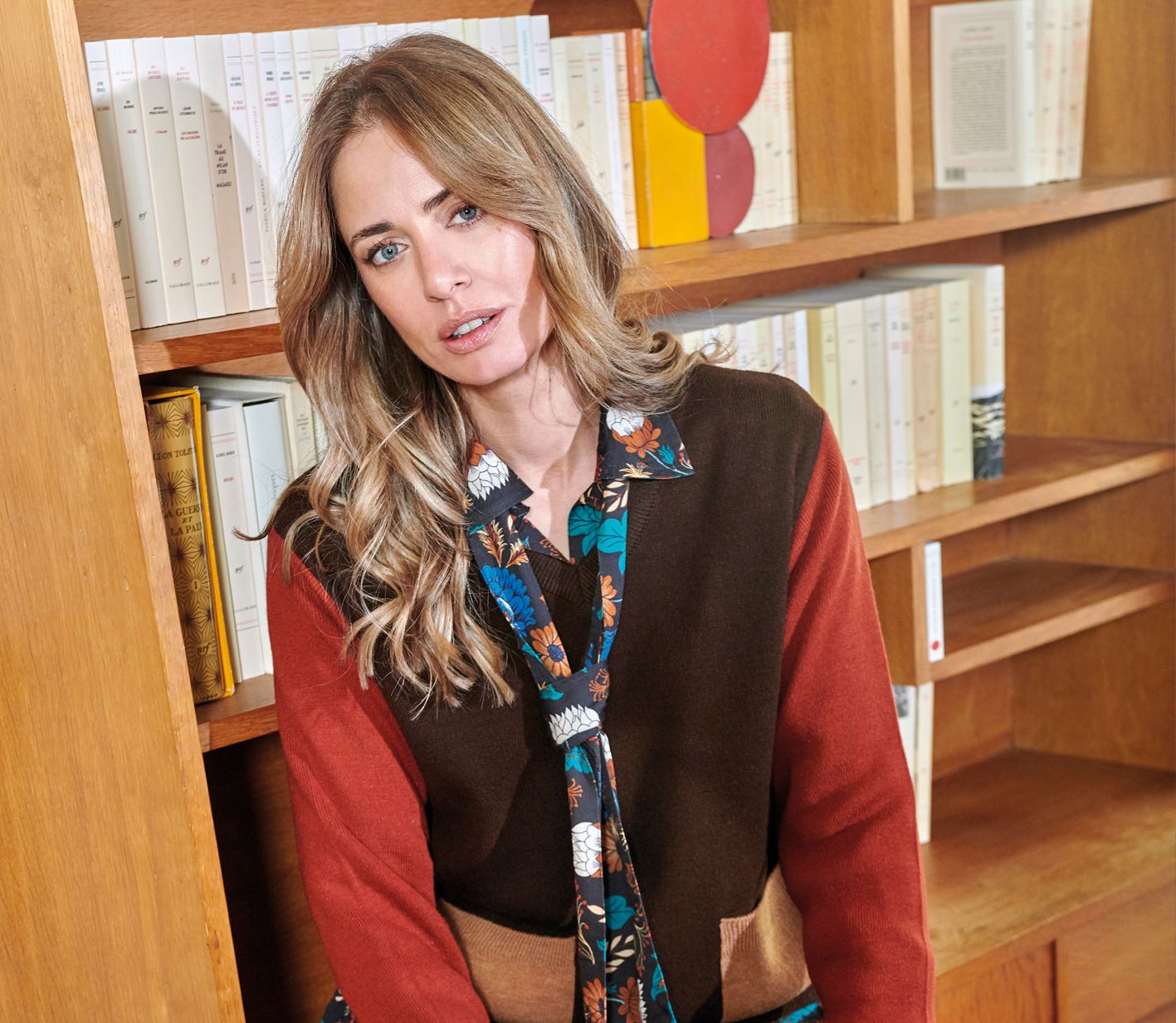 Femme assise dans une bibliothèque, portant une écharpe à motifs et un cardigan, regardant l’objectif.