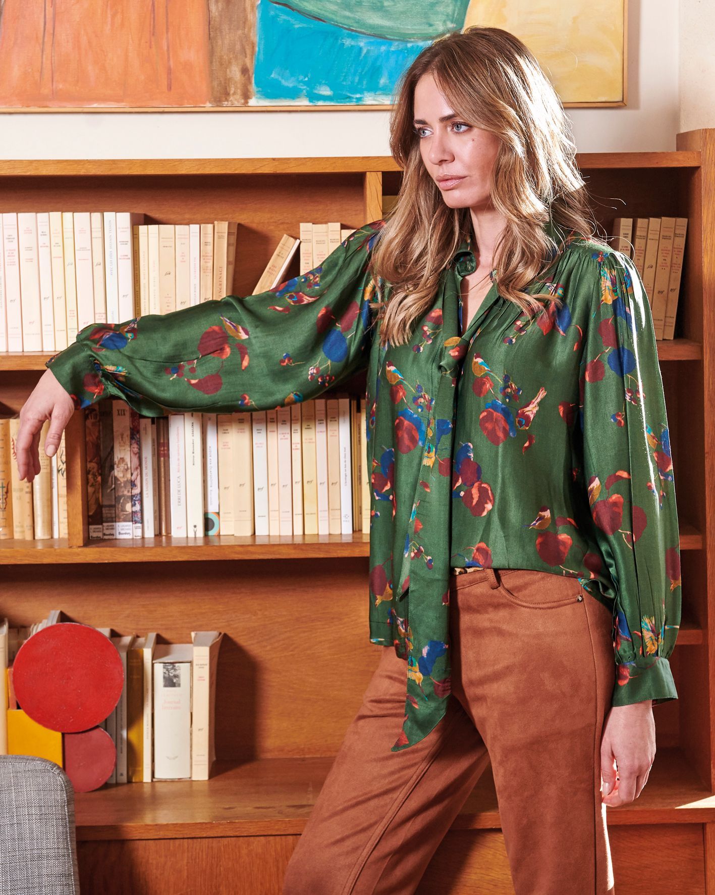Femme debout près d’une bibliothèque, vêtue d’un chemisier et d’un pantalon, dans une pièce.
