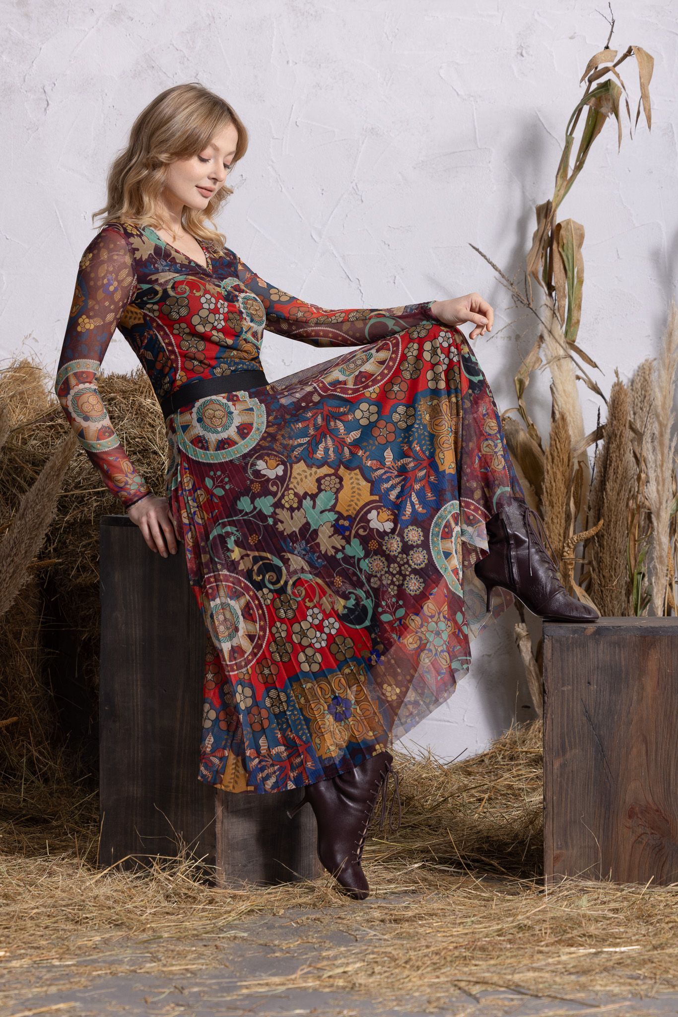 Femme en robe à fleurs et en bottes posant en extérieur à côté de bottes de foin et de hautes plantes séchées.