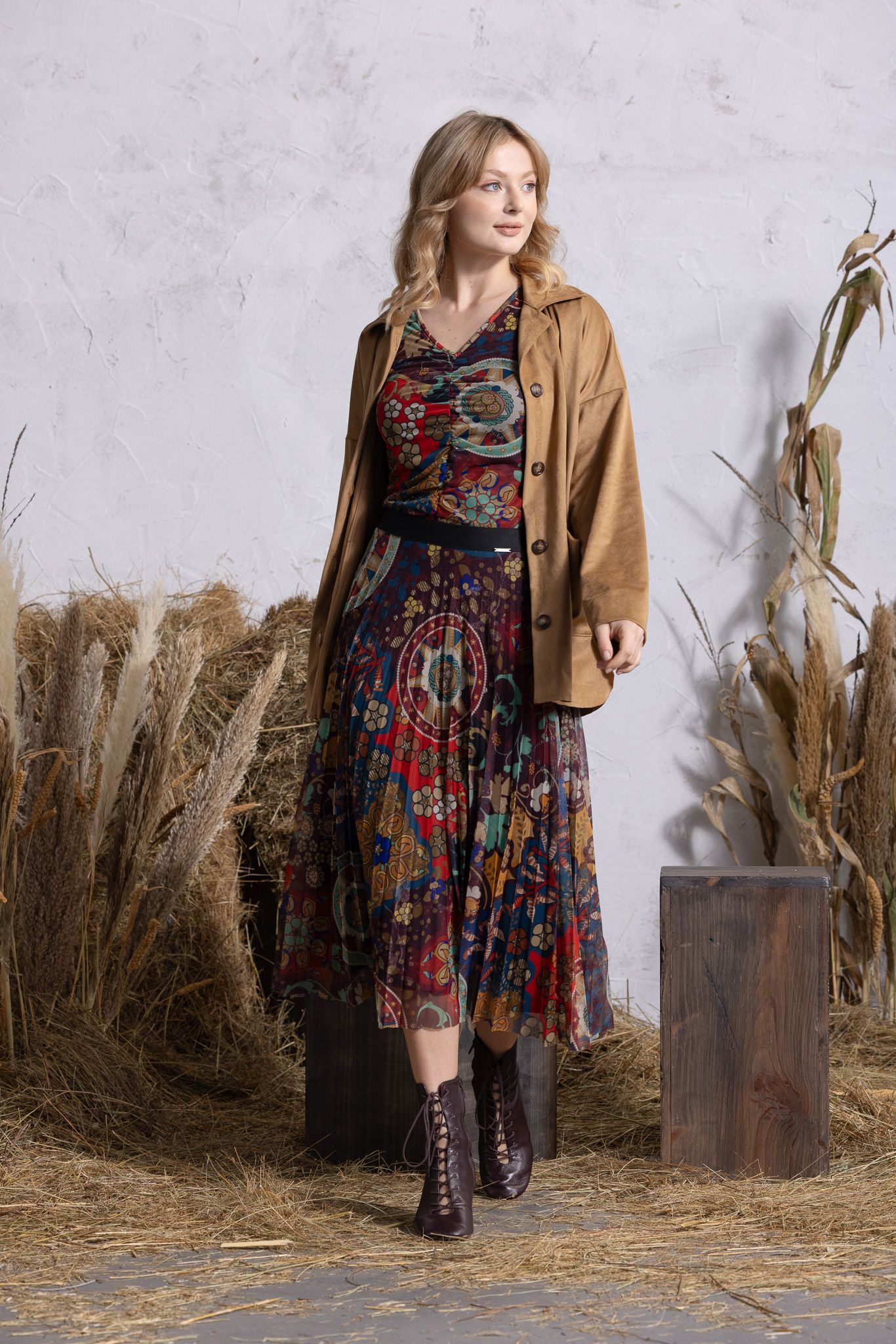 Femme en robe à fleurs et veste, debout dans un studio rustique avec des herbes séchées et un tabouret.