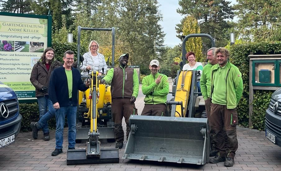 Eine Gruppe von Personen posiert mit Baumaschinen vor einem Gartenschild, im Hintergrund sind Bäume zu sehen.