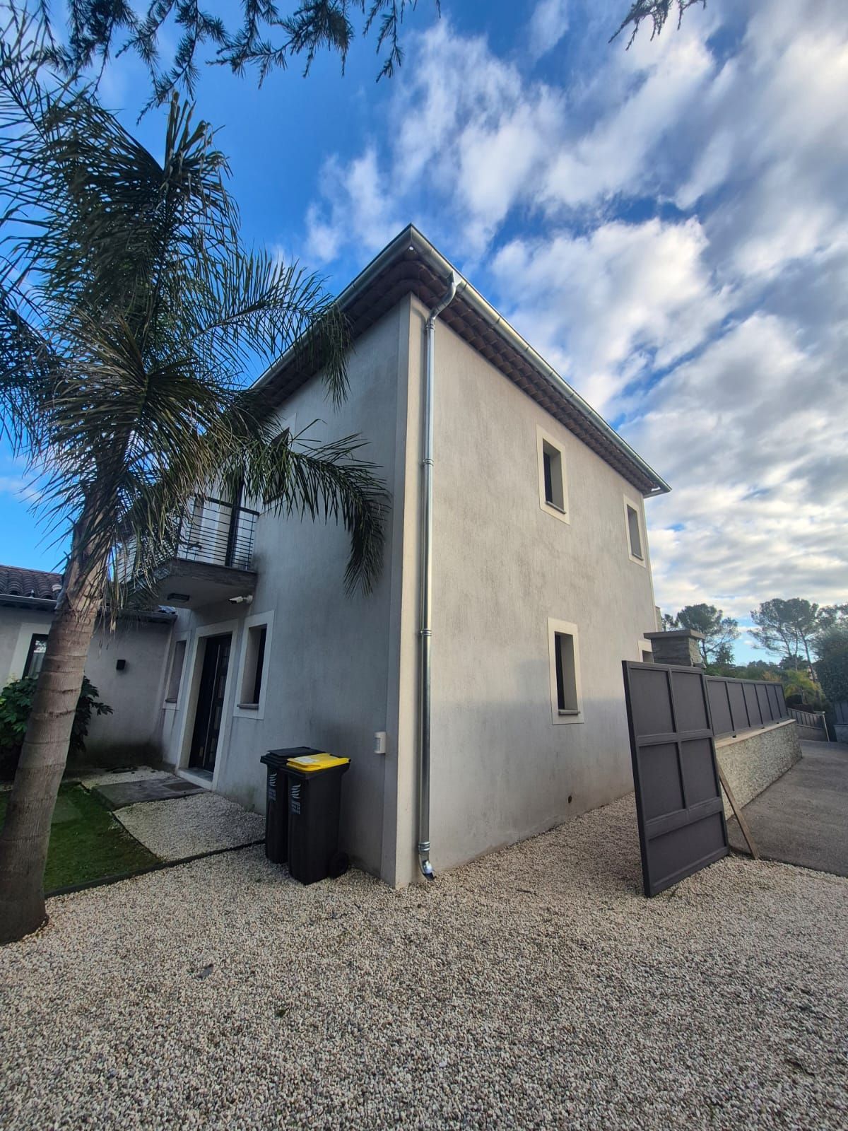 Maison en stuc de deux étages avec palmier, allée de gravier et portail partiellement visible par temps nuageux.