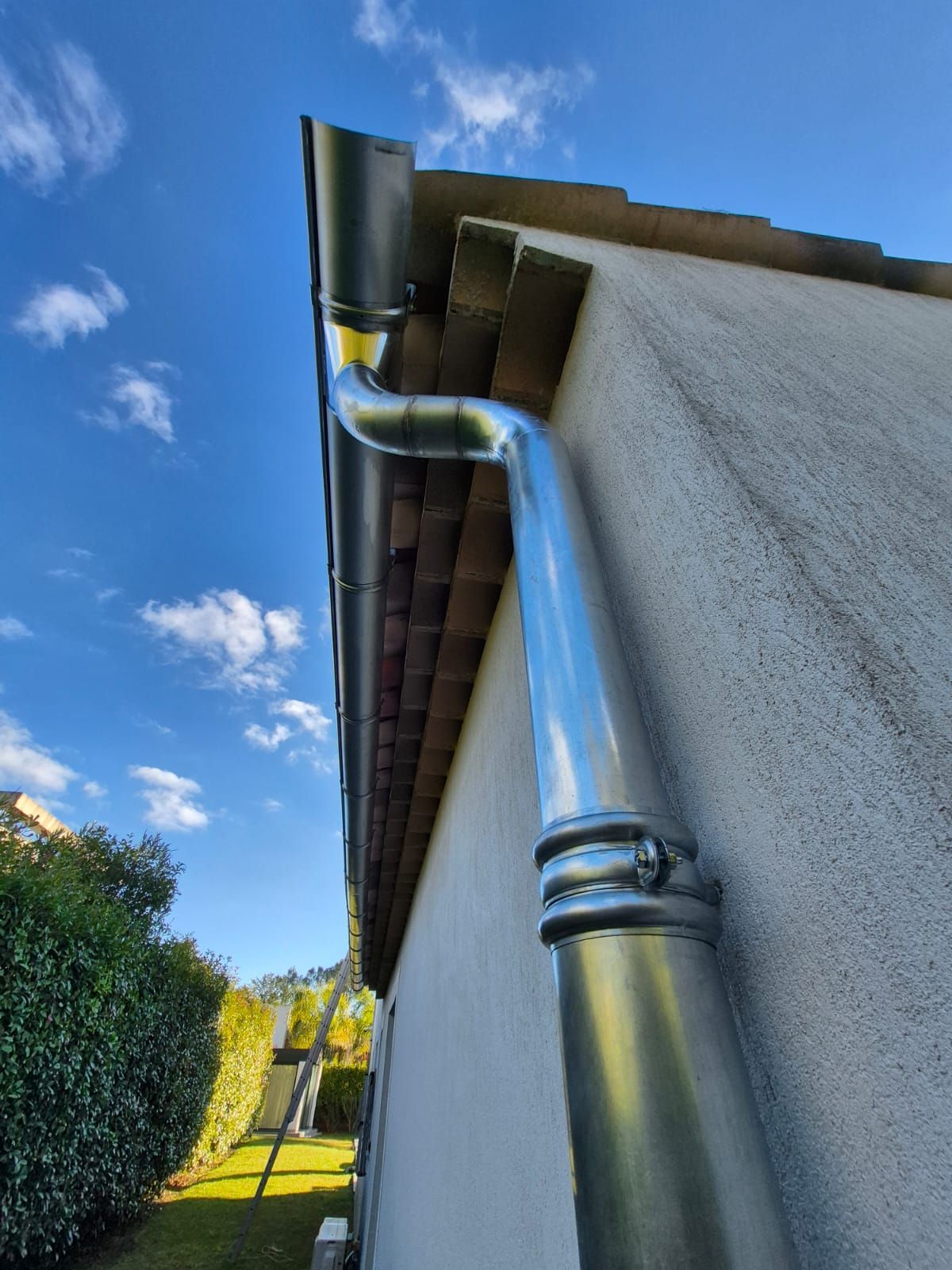 Nouvelles gouttières en métal argenté sur une maison avec un ciel bleu et un peu de verdure en arrière-plan.