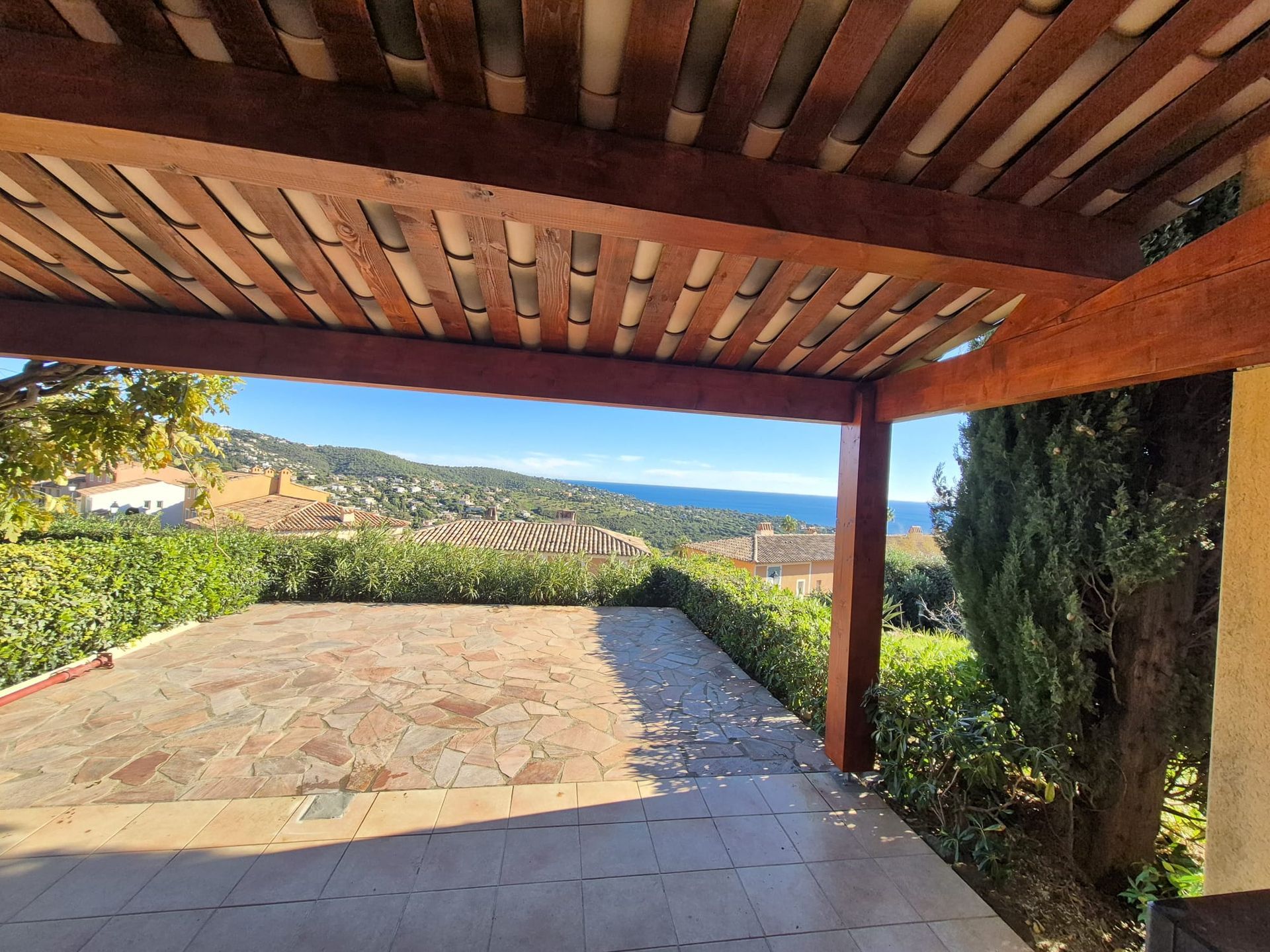 Pergola surplombant un paysage côtier ; bois brun, terrasse carrelée et vue sur la mer bleue.