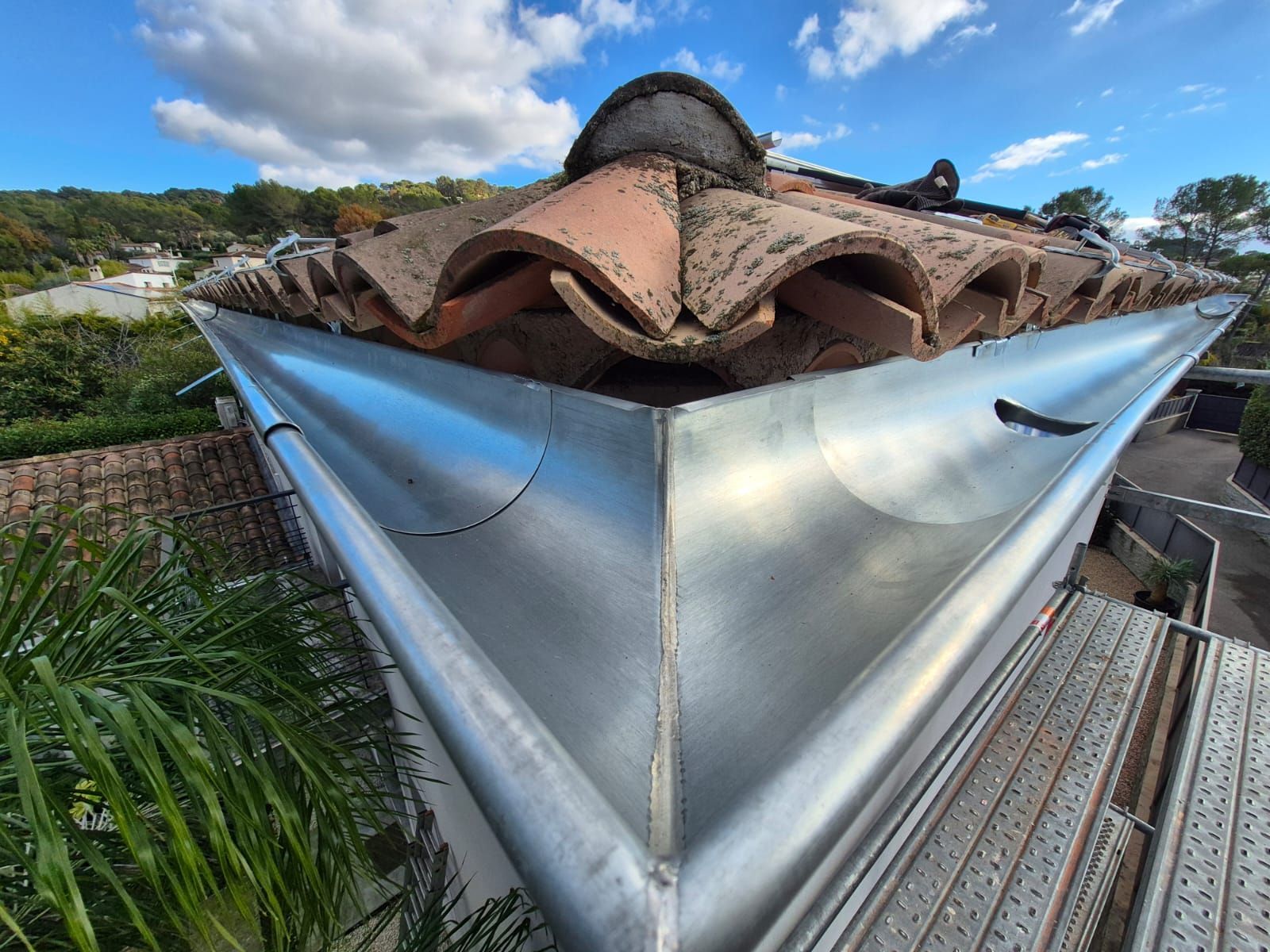 Nouvelle gouttière métallique installée à l'angle d'un toit en tuiles de terre cuite, sur fond de ciel bleu arboré.