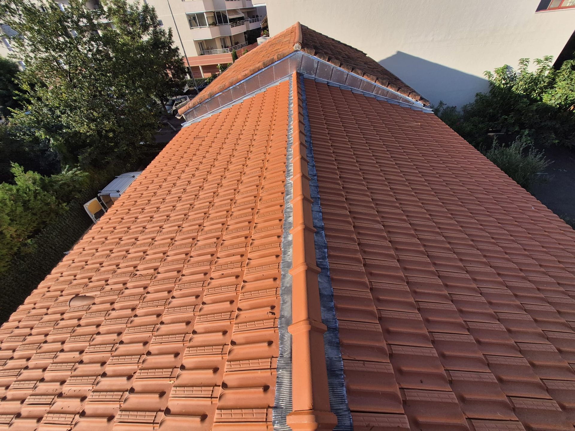 Vue d'un toit de tuiles en terre cuite avec une arête centrale, entouré d'arbres et d'un bâtiment en arrière-plan.