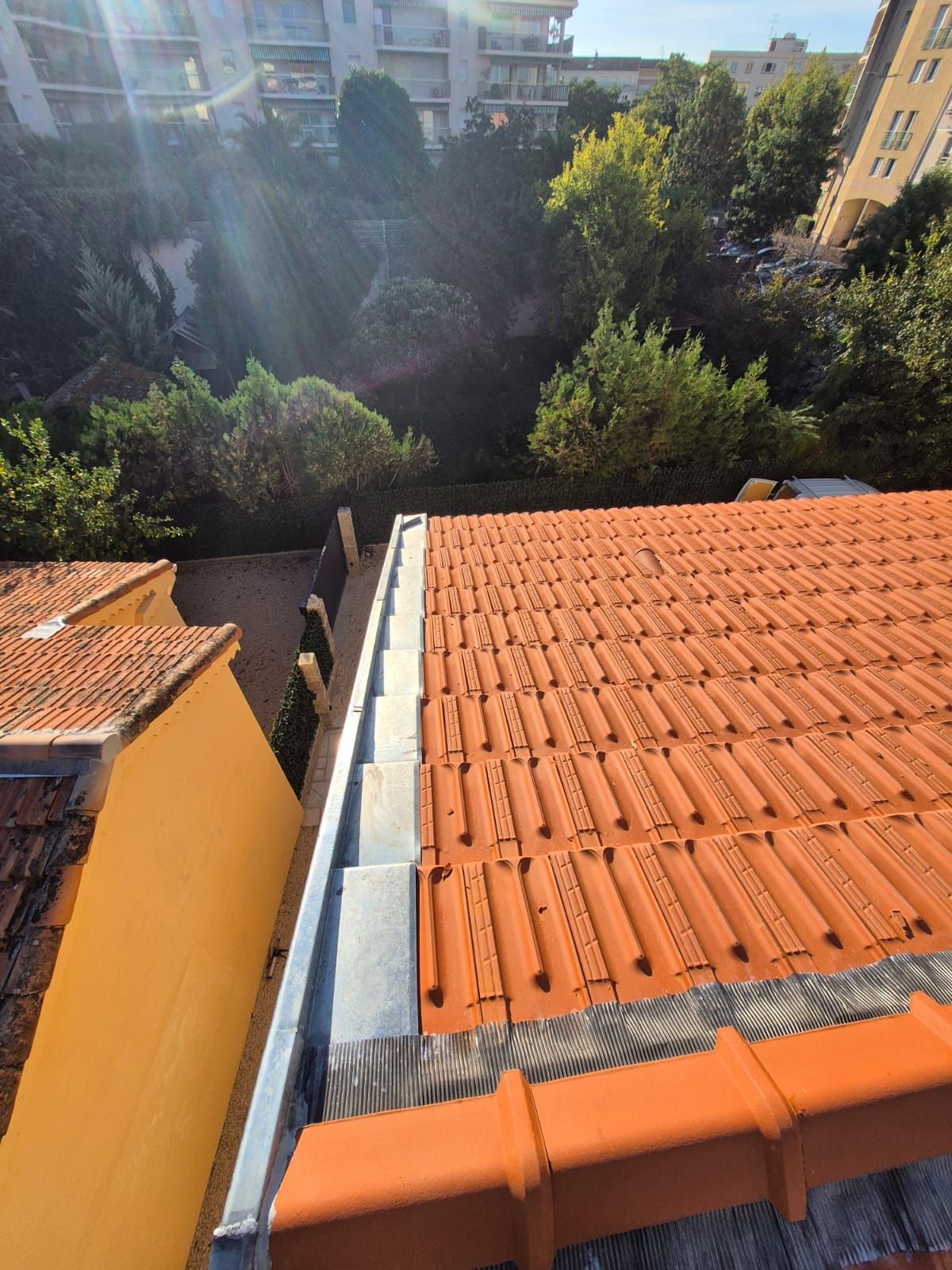 Toit de tuiles orange avec gouttière métallique, bâtiment jaune et arbres verts en arrière-plan.