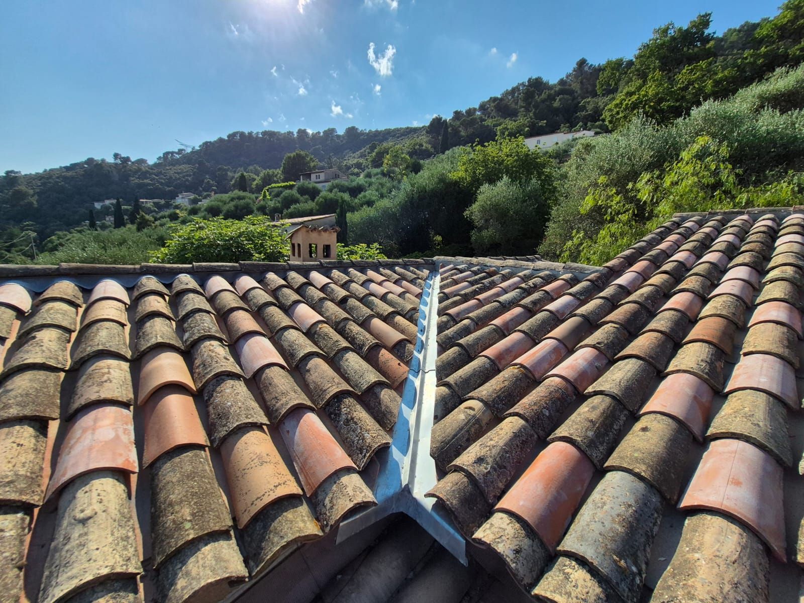 Toiture en tuiles de terre cuite avec solins métalliques, ensoleillée, vue sur une colline arborée et bâtie.