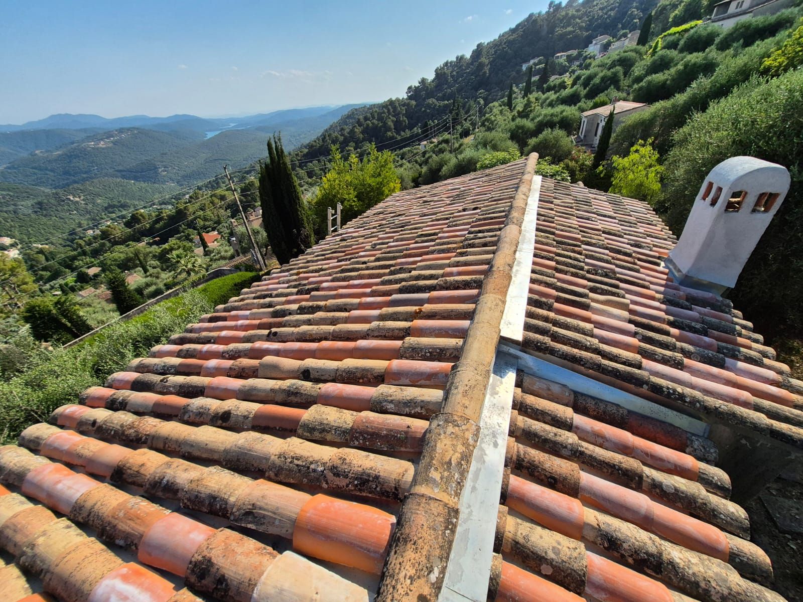 Toit de tuiles en terre cuite avec une cheminée donnant sur une vallée et des collines au loin.