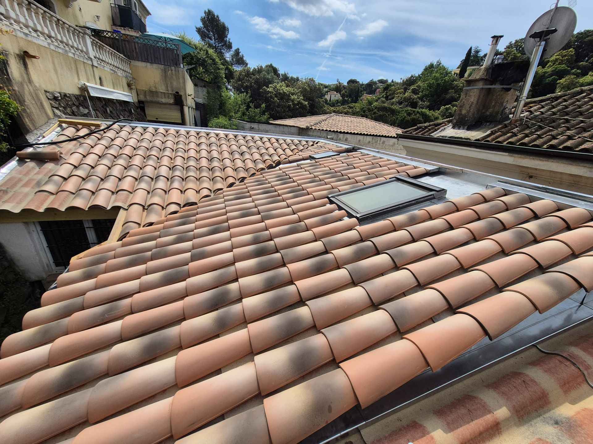 Toit en tuiles de terre cuite avec puits de lumière, bâtiments environnants et arbres sous un ciel bleu.