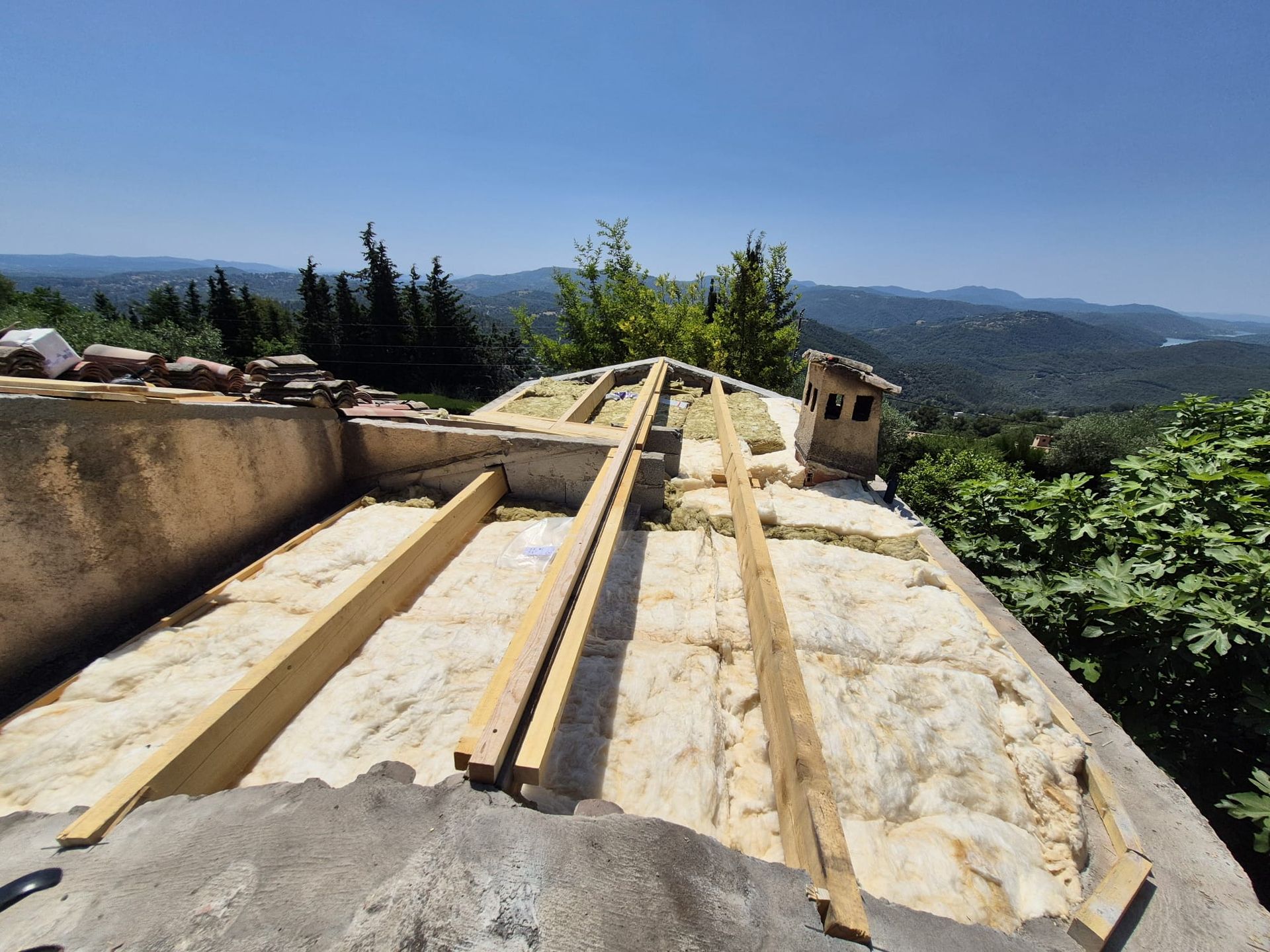 Toiture en construction, avec poutres en bois et isolation. Vue panoramique sur les arbres et les montagnes en arrière-plan.