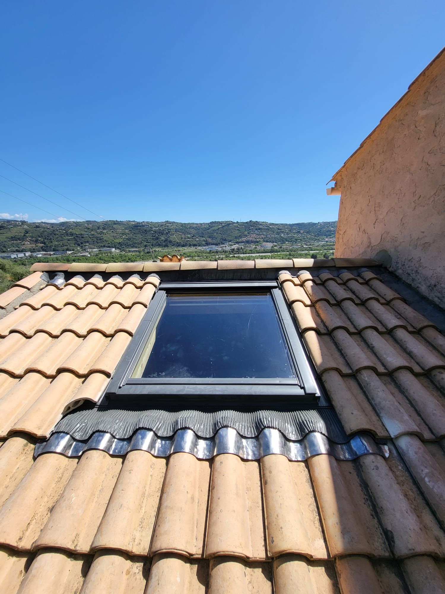 Puits de lumière sur un toit de tuiles offrant une vue sur un paysage verdoyant sous un ciel bleu.