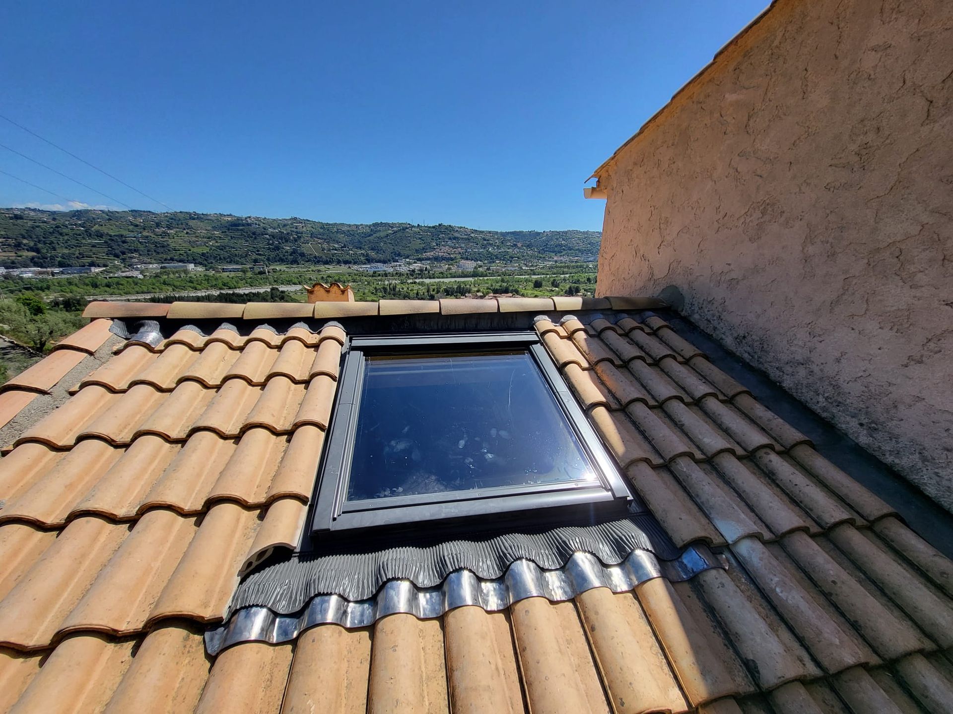 Puits de lumière sur un toit de tuiles en terre cuite offrant une vue sur une ville au loin sous un ciel bleu.