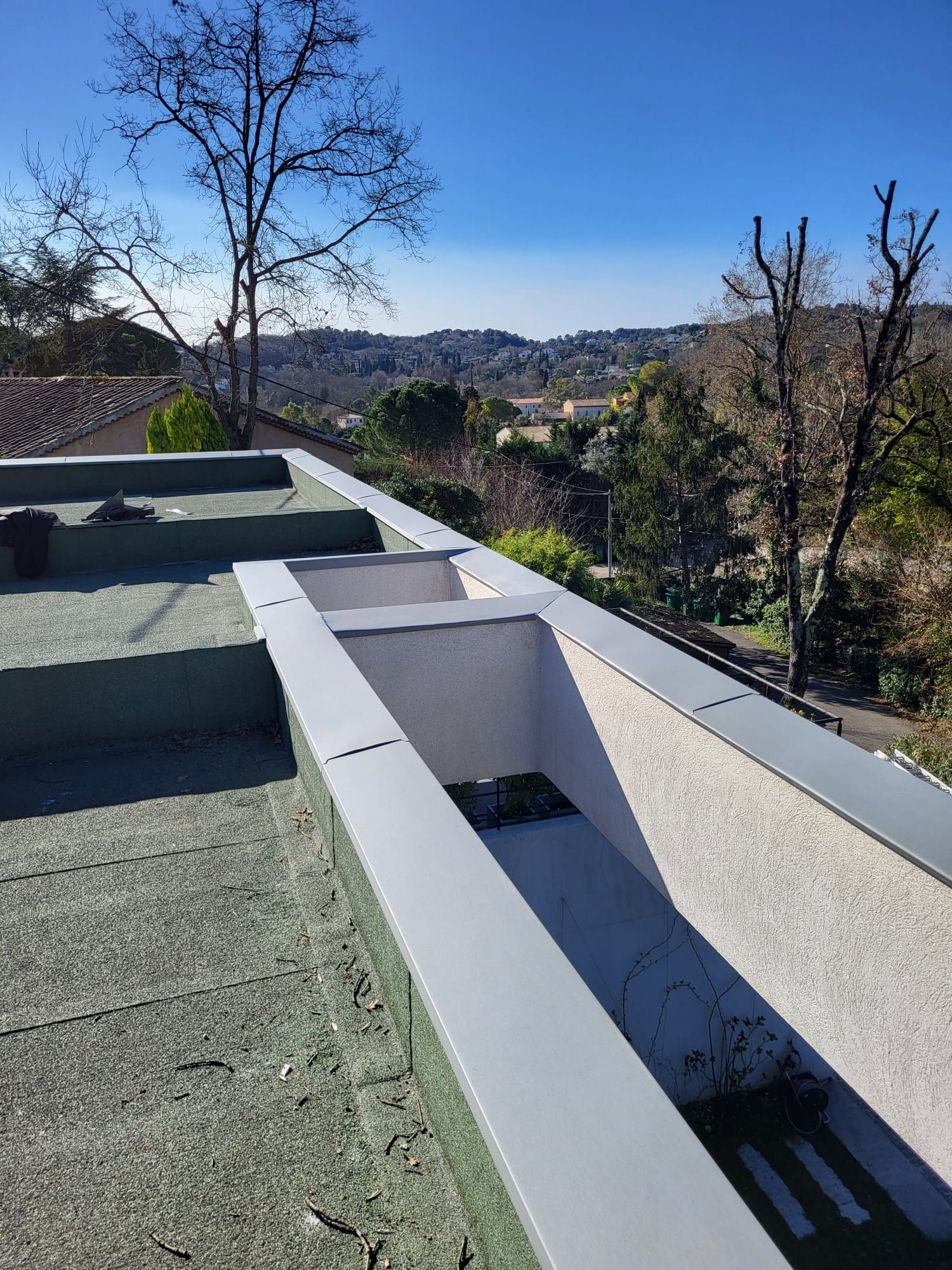 Toit plat avec une gouttière rectangulaire, des finitions grises et une vue sur les arbres et les collines.