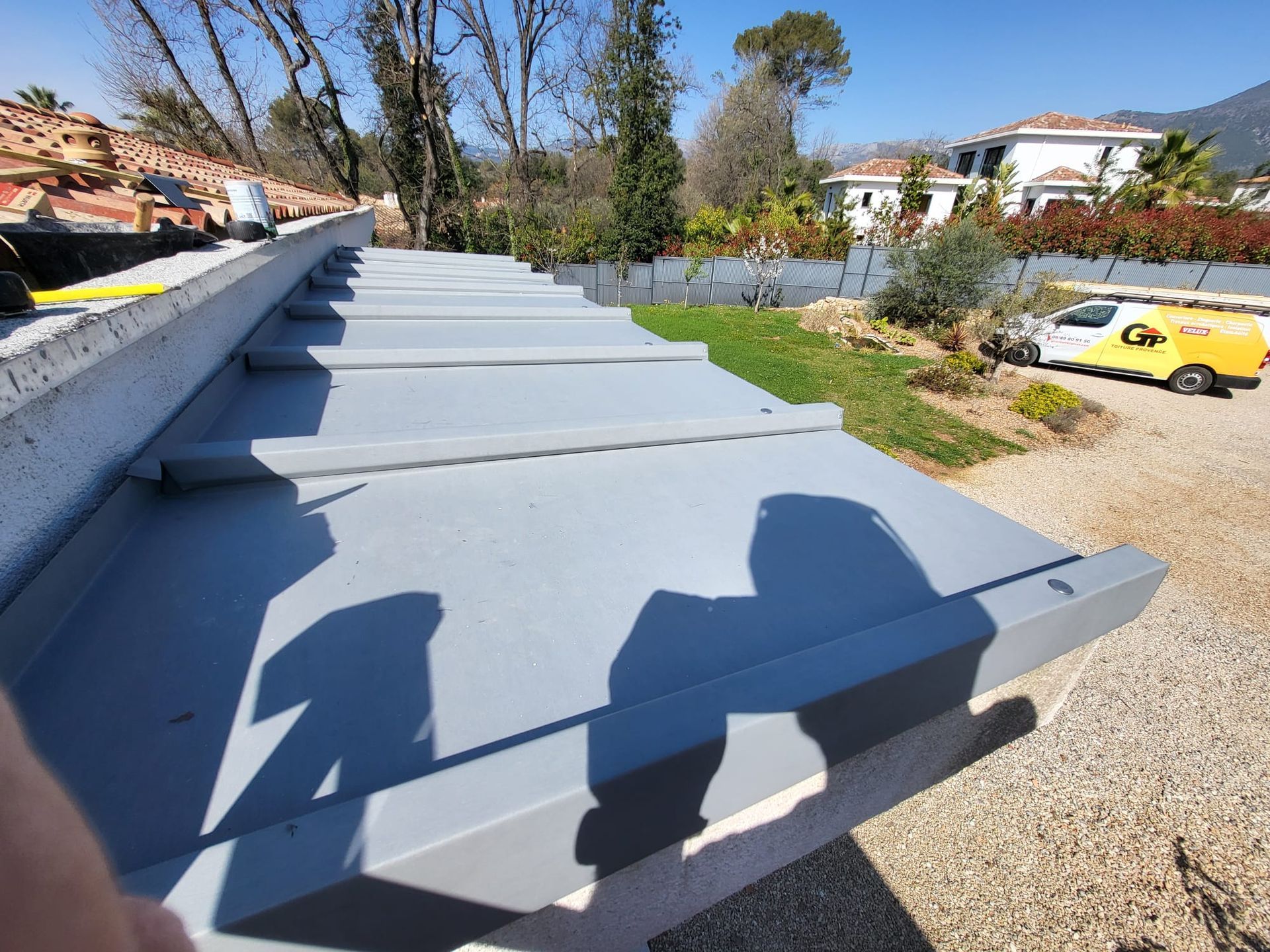 Toiture métallique grise avec ombres, gravier, herbe, arbres et bâtiments en arrière-plan par une journée ensoleillée.