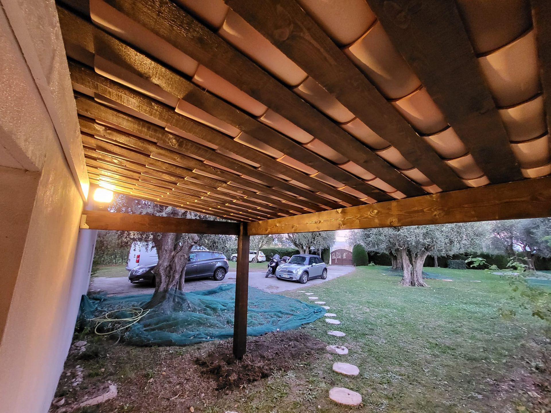 Pergola en bois avec toit de tuiles en terre cuite, donnant sur une cour avec des voitures garées et des oliviers.