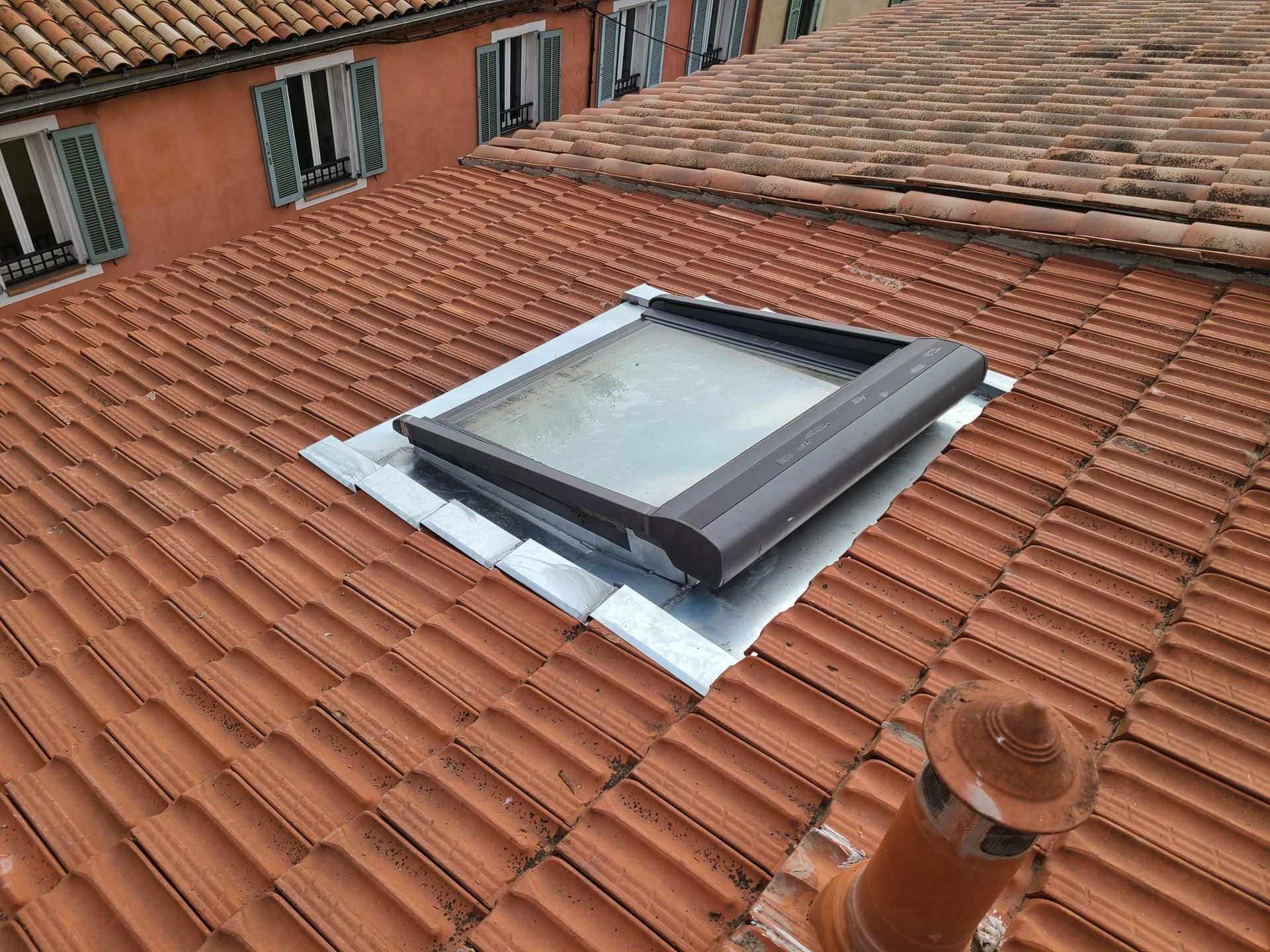 Puits de lumière sur un toit de tuiles en terre cuite, avec une solin métallique. Cheminée au premier plan.