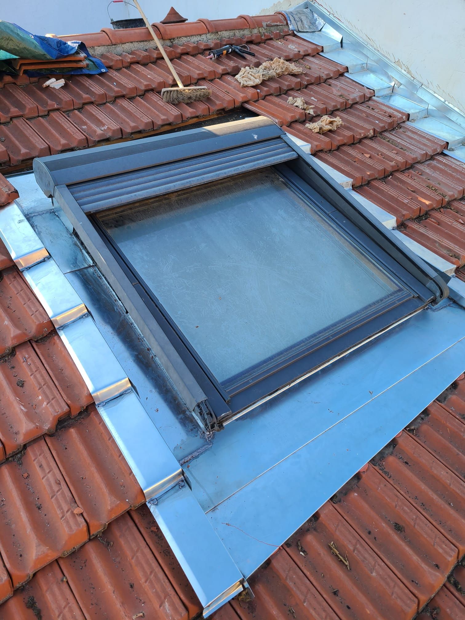 Puits de lumière sur un toit de tuiles rouges, entouré de solins argentés. 