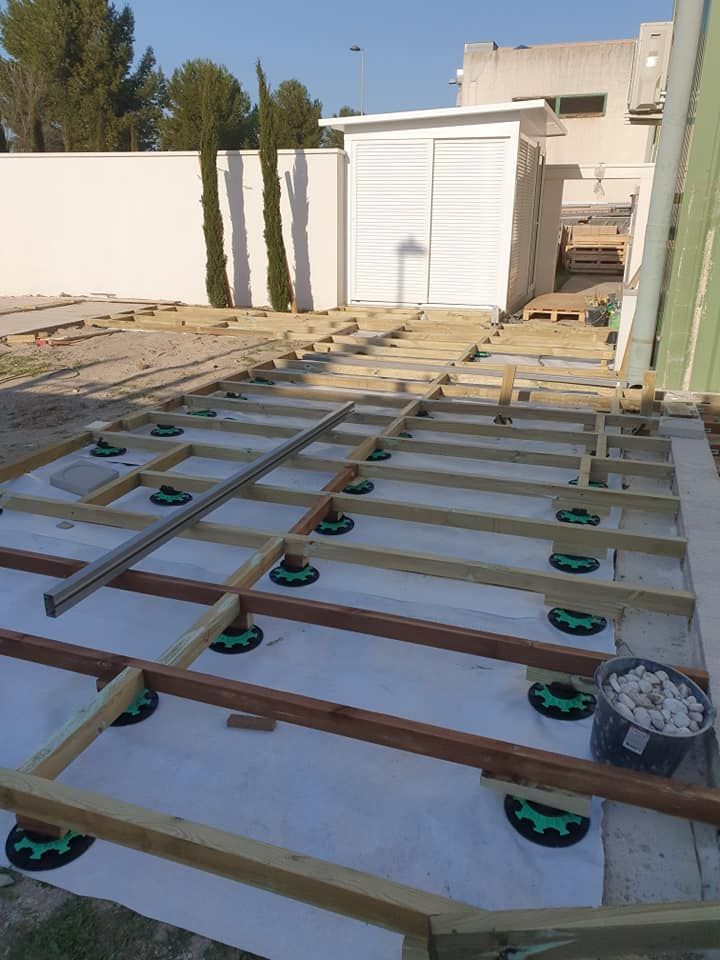 Terrasse surélevée en bois dans un jardin ensoleillé, avec un abri de jardin blanc et une clôture en arrière-plan