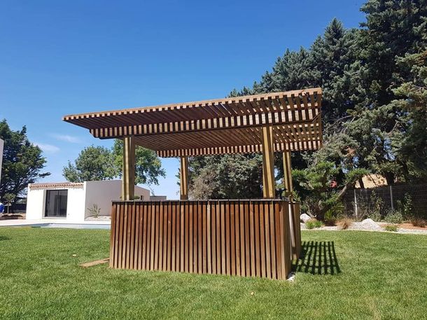 Kiosque extérieur en bois avec toit à claire-voie et comptoir dans un parc verdoyant sous un ciel bleu azur