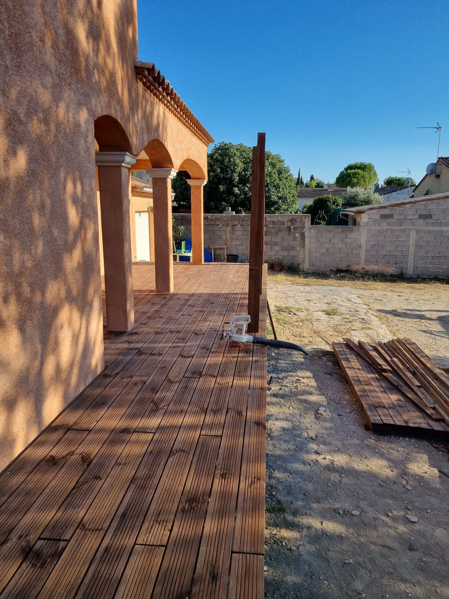 Patio ensoleillé jouxtant une maison en stuc, avec une terrasse en bois, des piliers et une cour clôturée