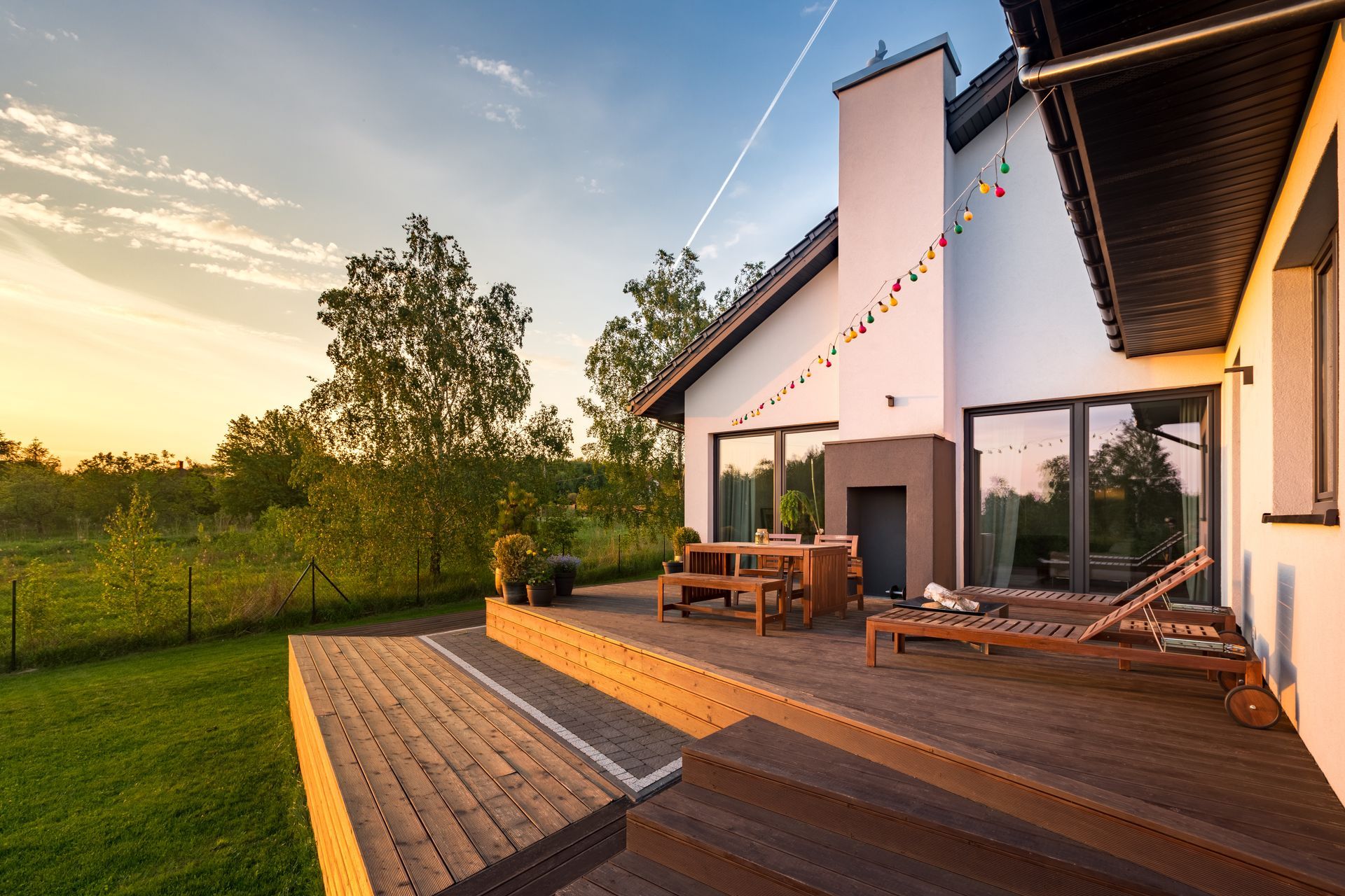 Terrasse en bois ensoleillée avec un ensemble de repas extérieur à côté d'une maison blanche, donnant sur un champ verdoyant au coucher du soleil
