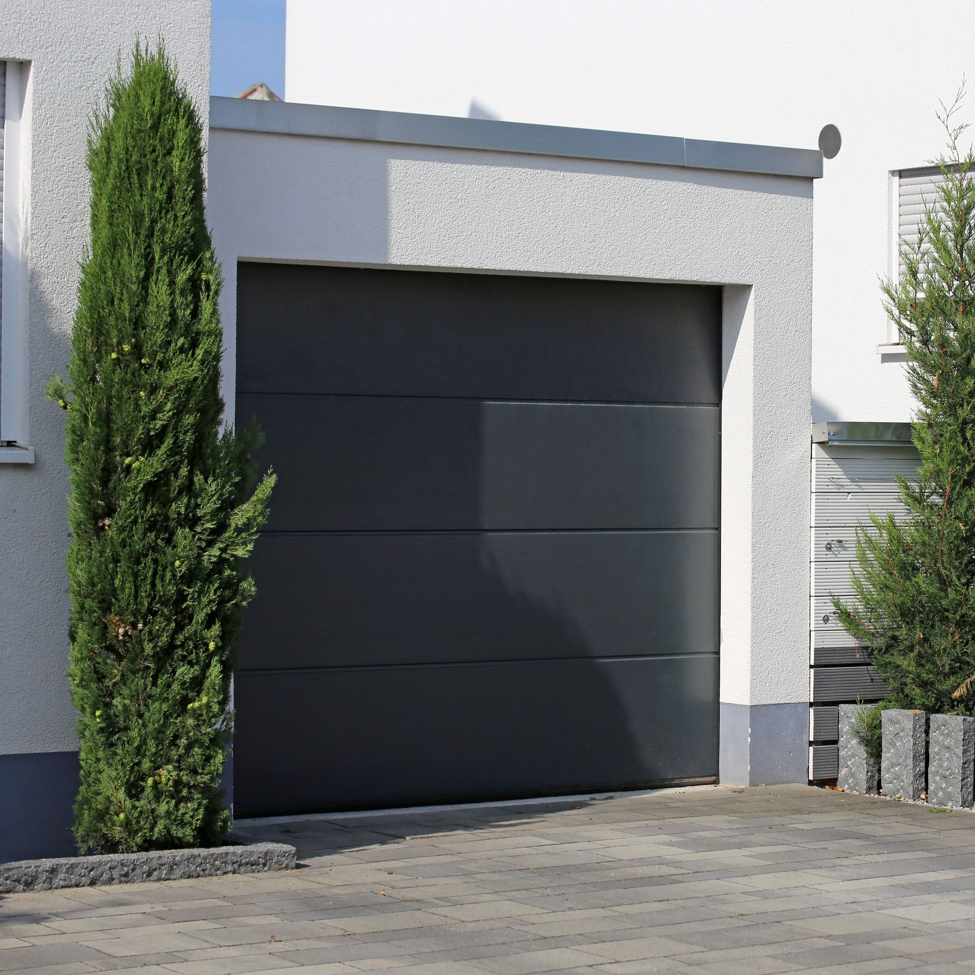 Extérieur de maison moderne avec une porte de garage noire et de grands arbres étroits bordant une allée pavée