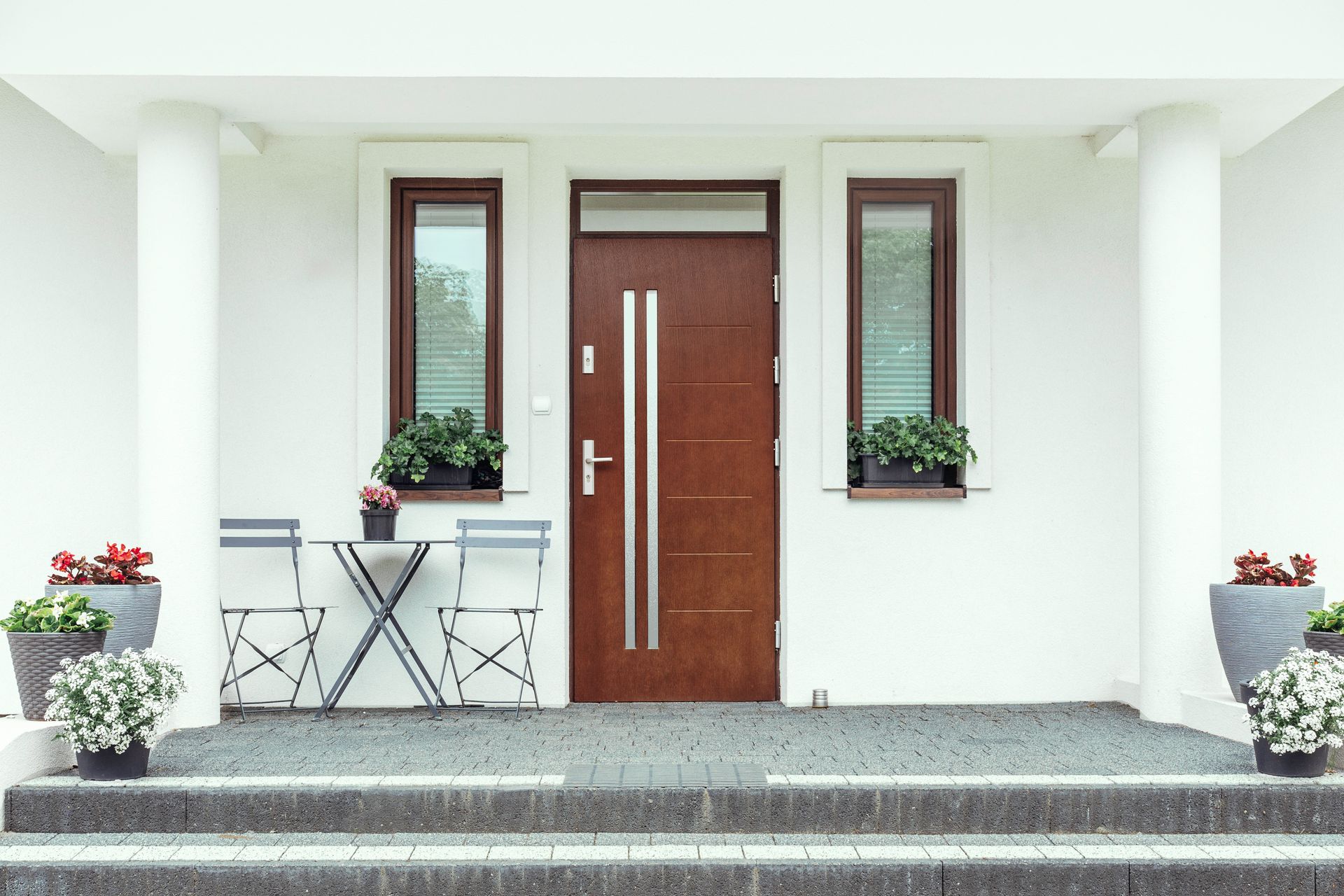 Porche d'entrée avec porte marron, façade blanche, plantes en pot et petite table et chaises de café