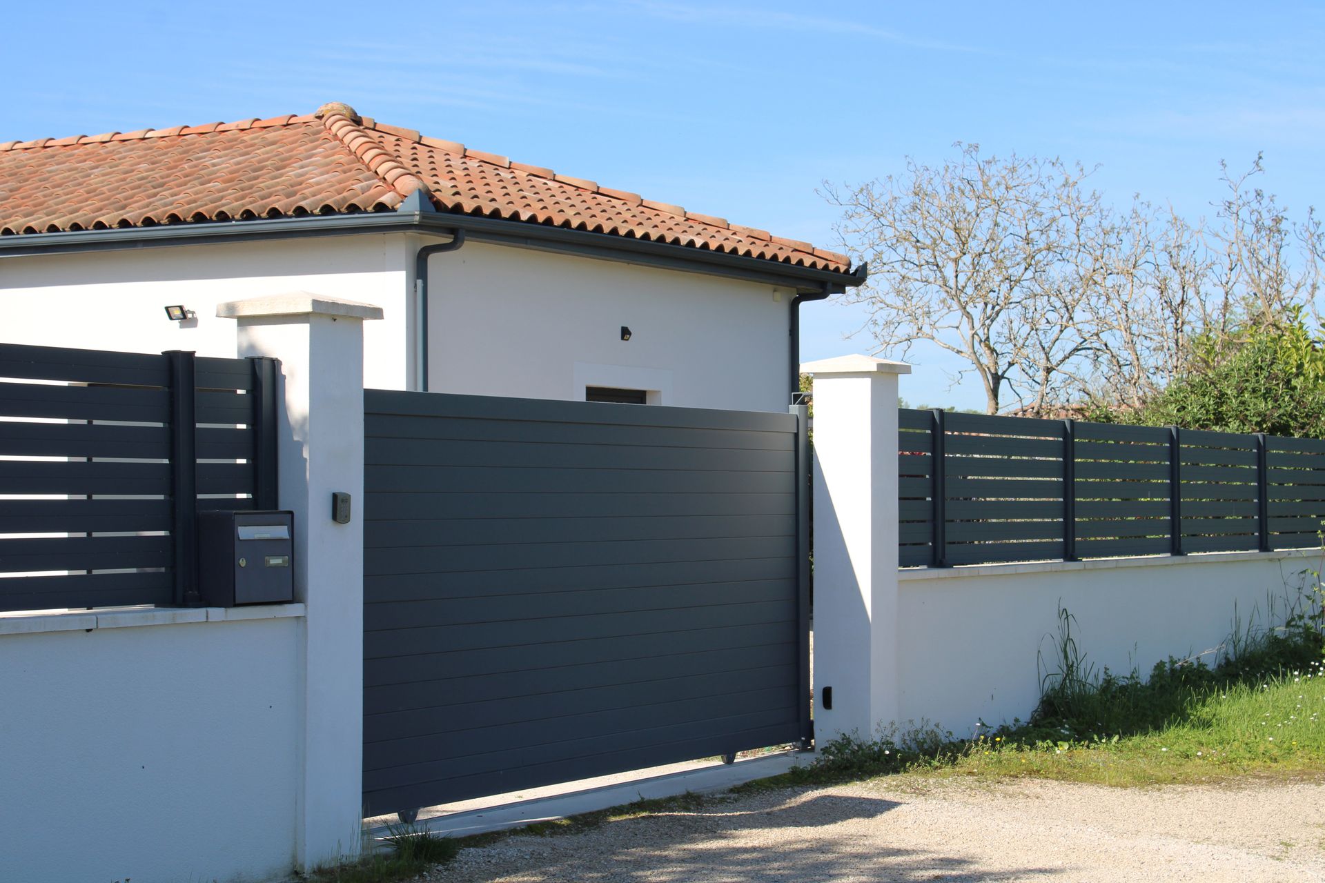 Maison blanche au toit de tuiles rouges et au portail gris foncé, derrière un mur blanc, par une journée ensoleillée