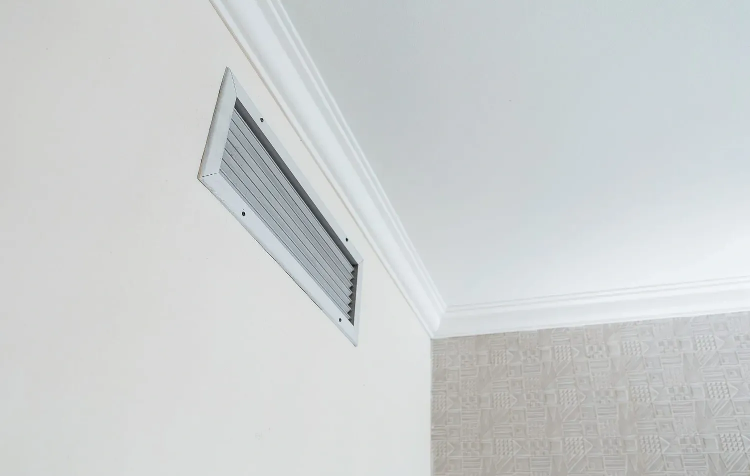Una rejilla de ventilación rectangular de metal está montada en una pared blanca cerca de un techo blanco.