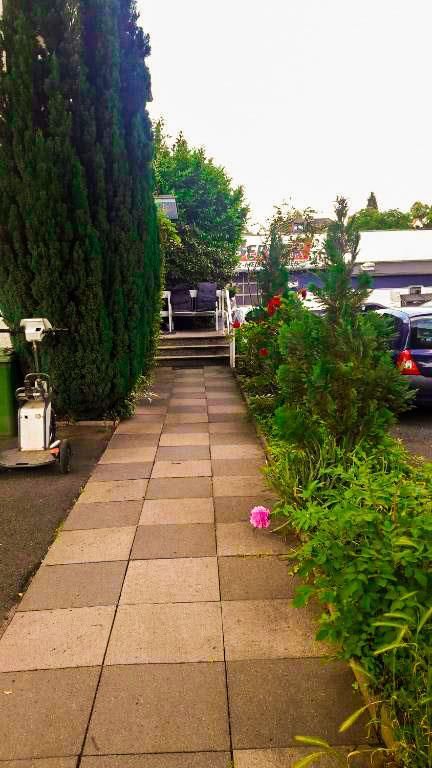 Ein gemauerter Gehweg führt zu einem von Bäumen und Büschen umgebenen Haus.