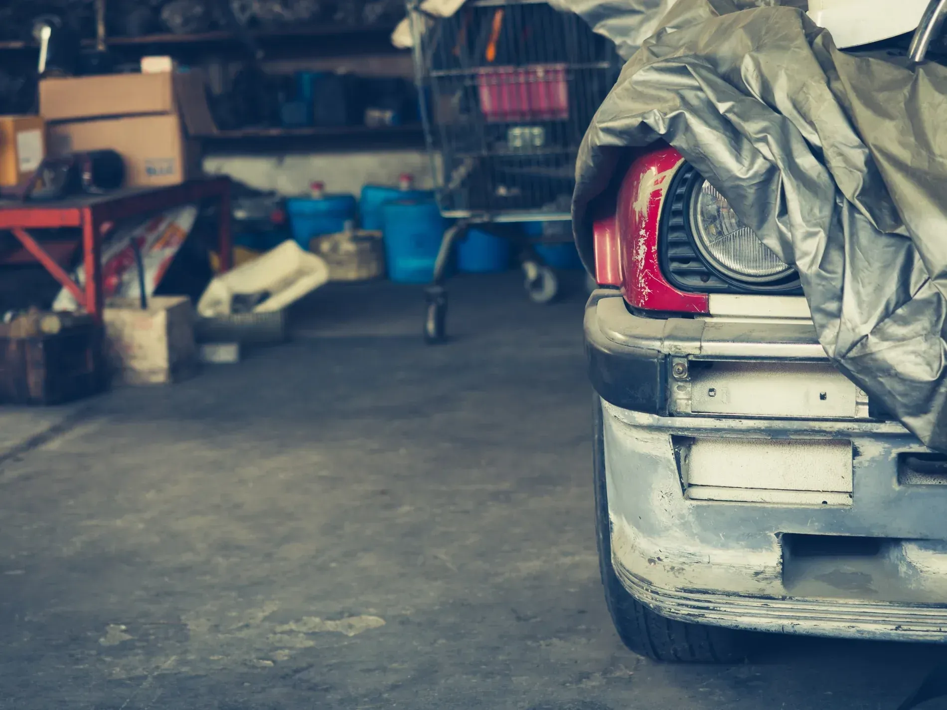 Coche plateado parcialmente cubierto en garaje, faro visible, con fondo desordenado.