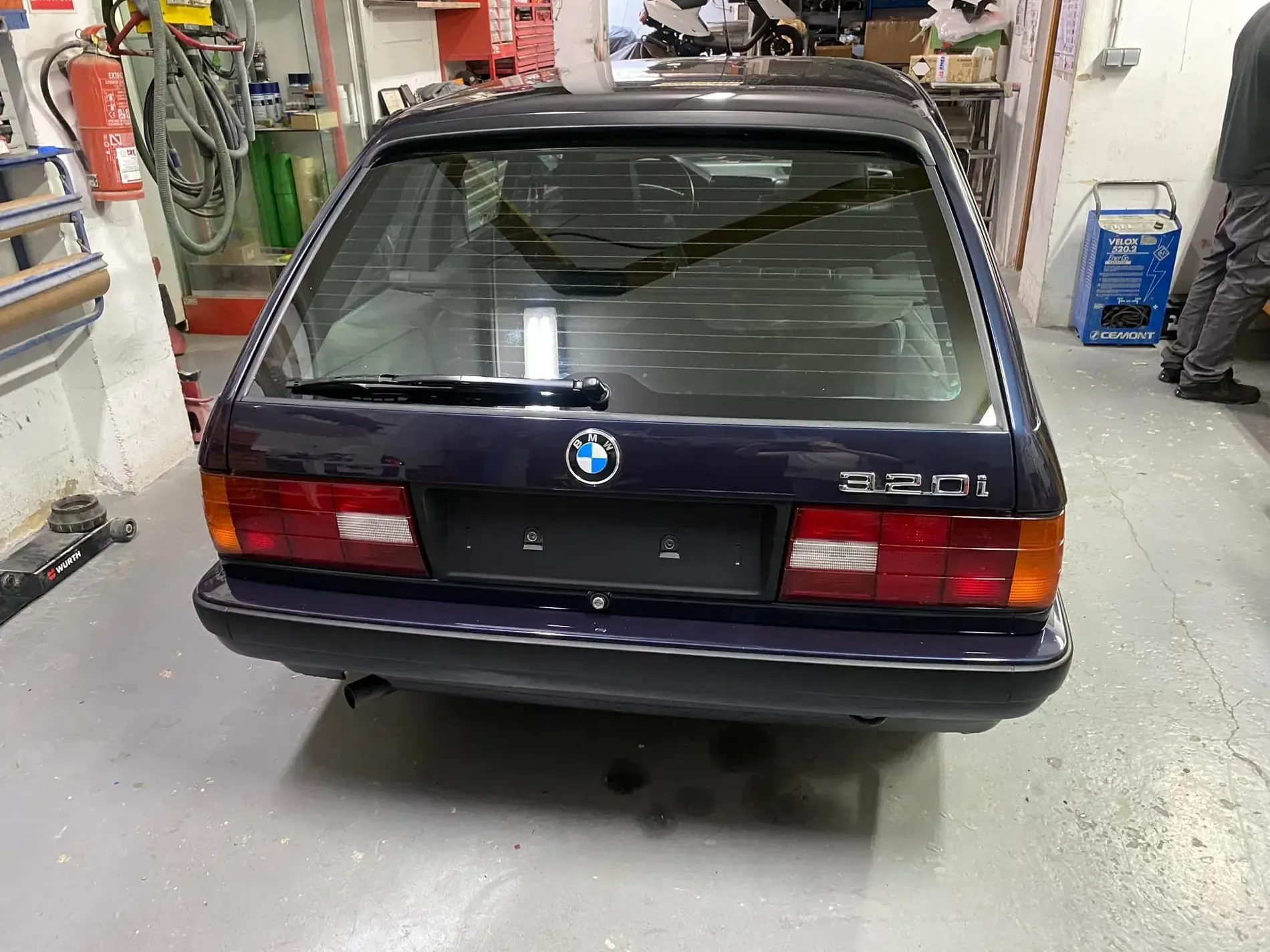 Un BMW 325i Touring azul oscuro estacionado en un garaje. Vista trasera, luces traseras visibles.