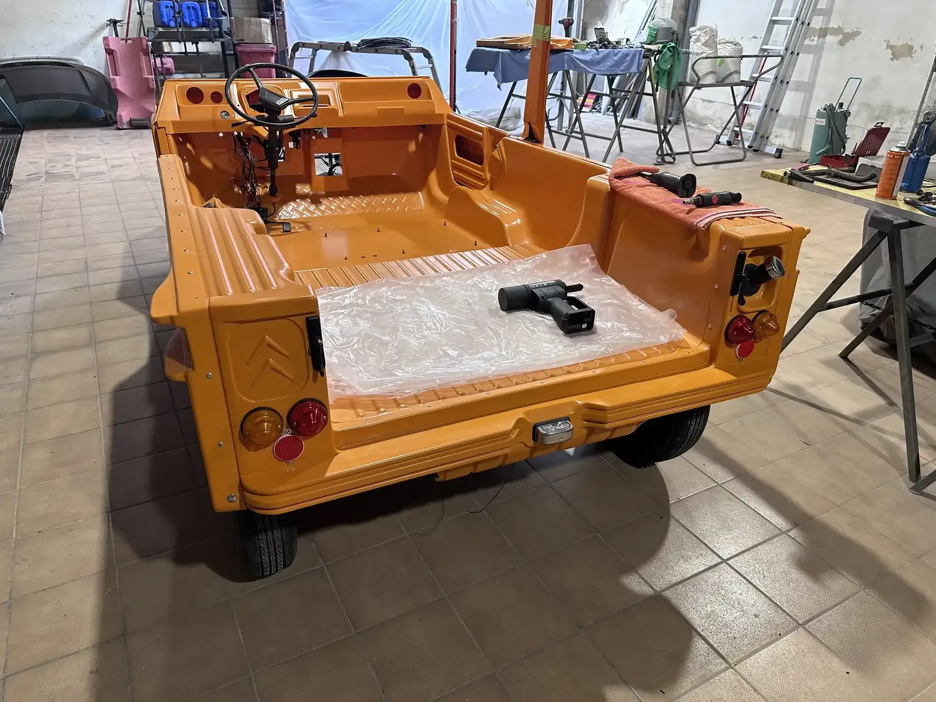 Vehículo utilitario abierto de color naranja en proceso de ensamblaje en un taller, vista trasera, con luces traseras.