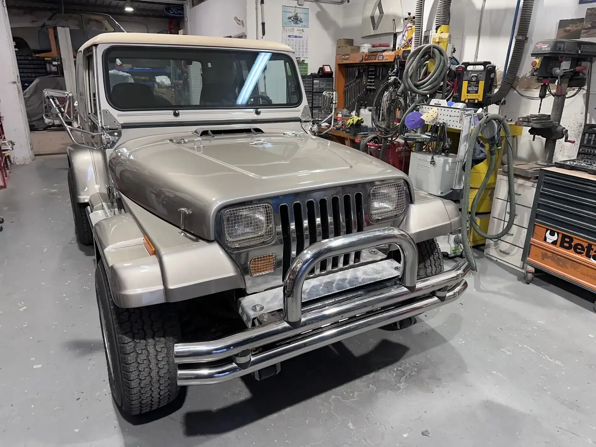 Jeep Wrangler de color tostado en un garaje con herramientas y equipos visibles.