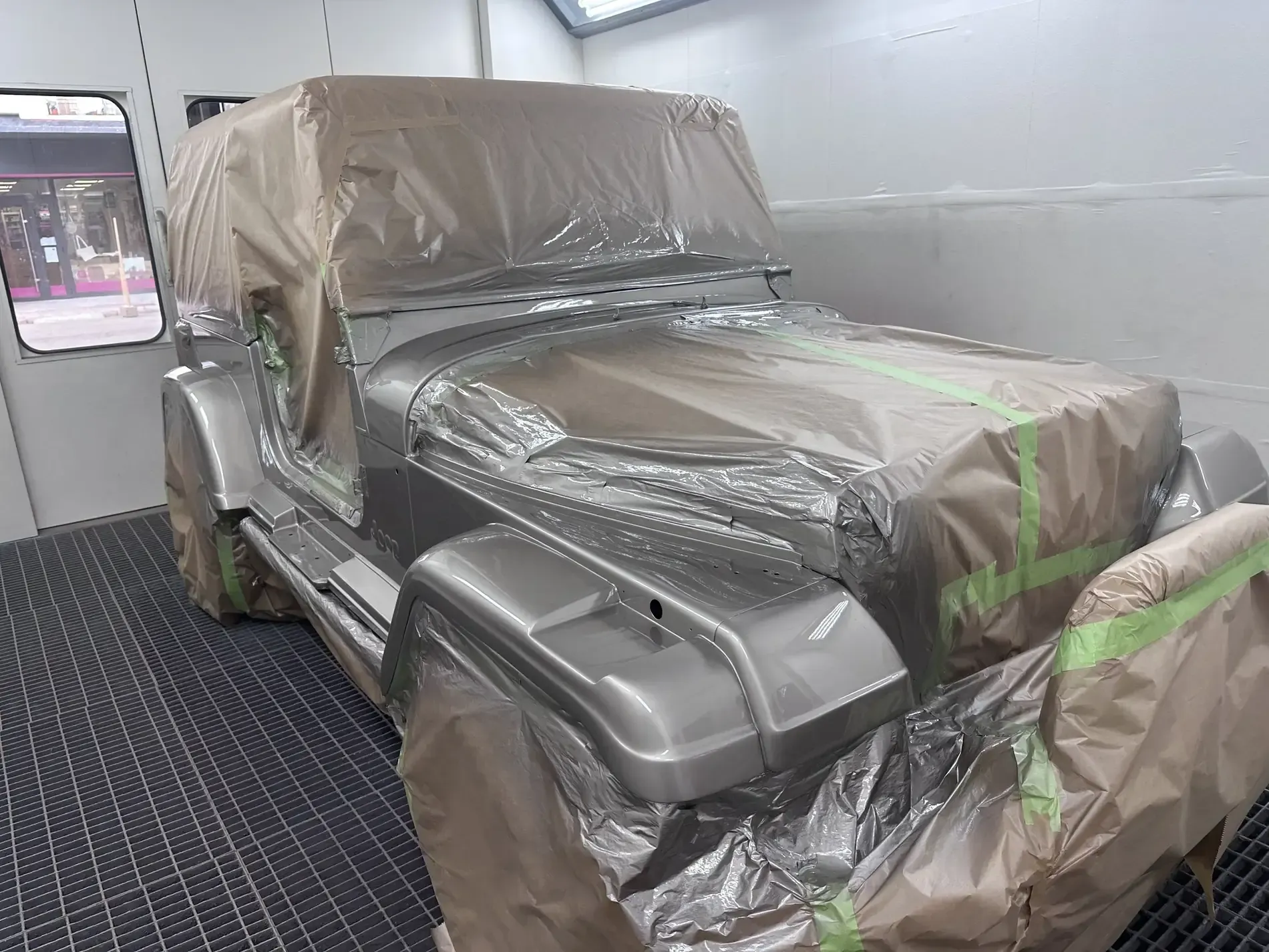 Un Jeep Wrangler gris, parcialmente cubierto con plástico y cinta, dentro de una cabina de pintura.