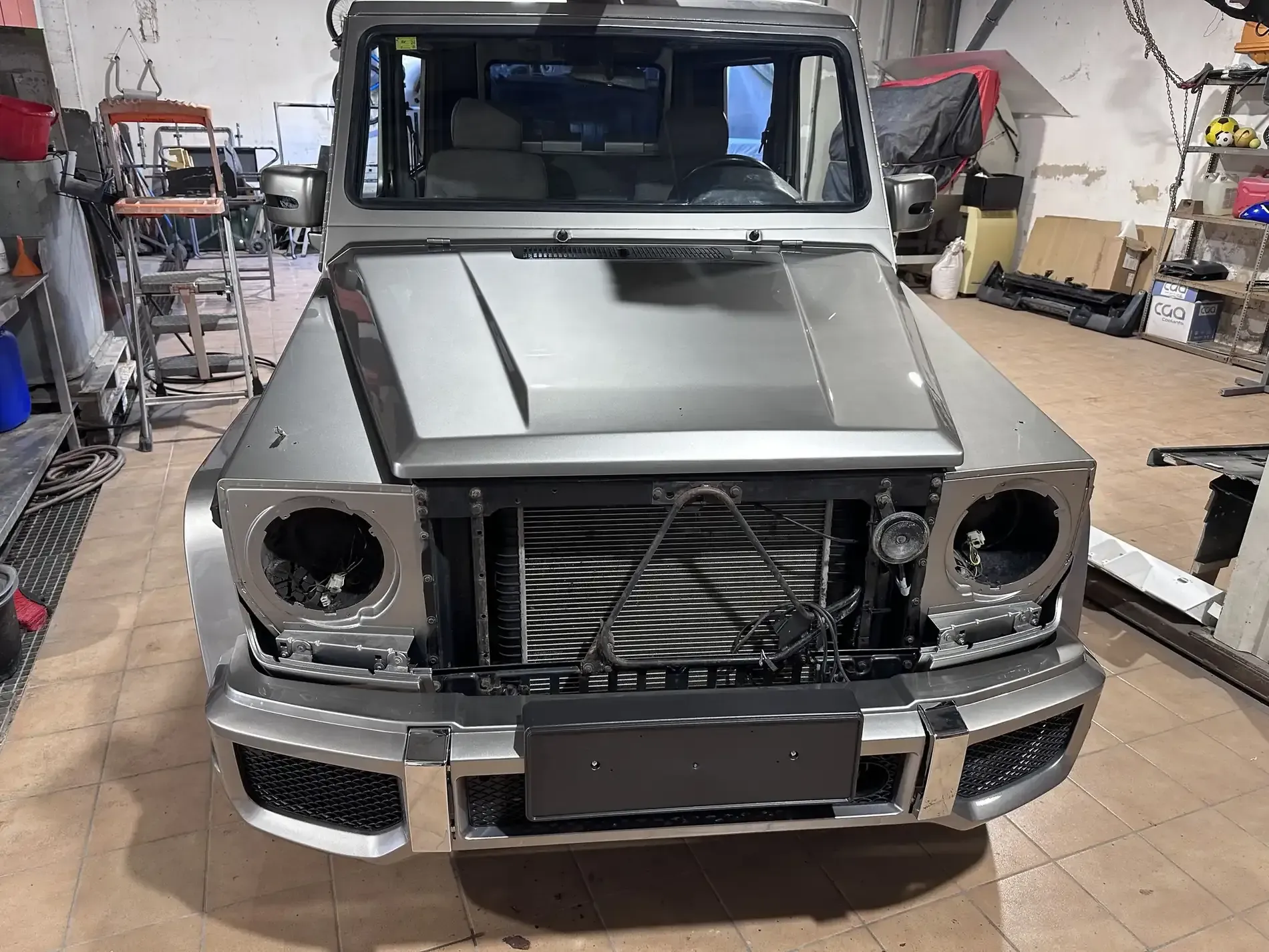 SUV Mercedes-Benz Clase G color plata en proceso de restauración en taller; vista frontal con faros faltantes.