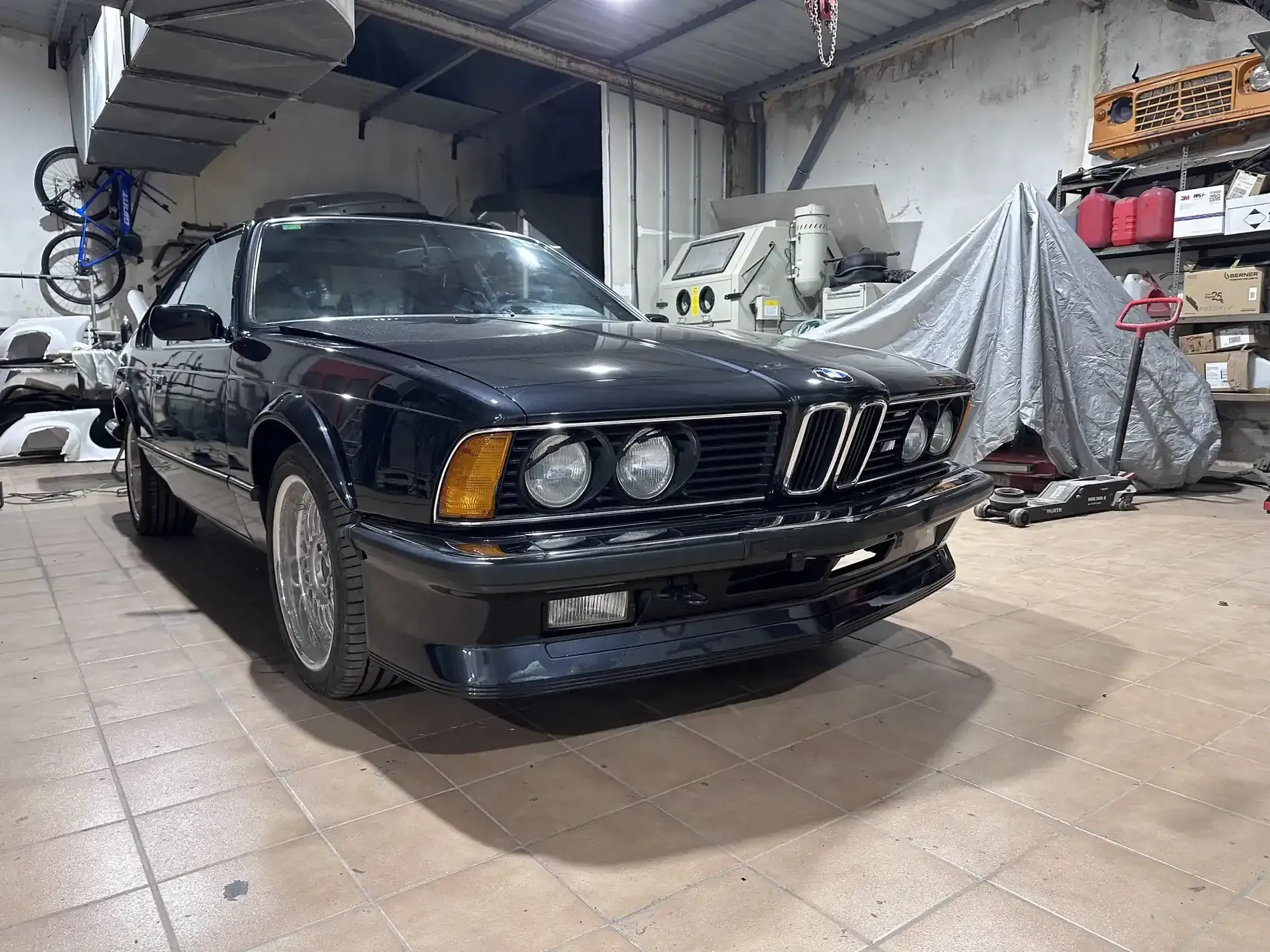 Coche BMW negro dentro de un garaje, con algunas piezas de automóvil y herramientas visibles.