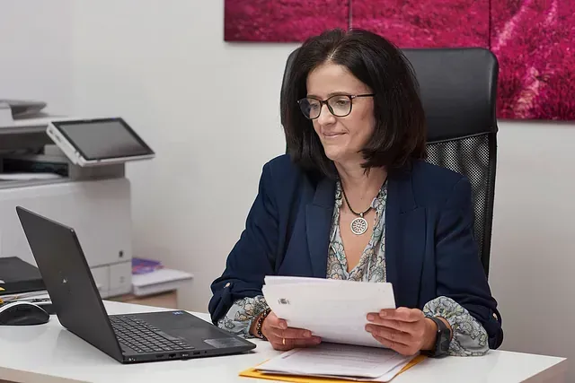 Mujer con chaqueta azul marino leyendo documentos en un escritorio con un portátil en una oficina.