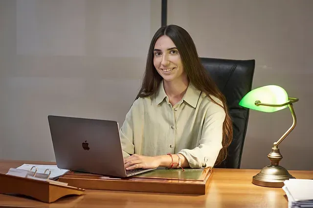 Mujer sentada en un escritorio con una computadora portátil y una lámpara verde, sonriendo en un entorno de oficina.