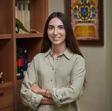 Mujer de pie con los brazos cruzados en una oficina, con estanterías y un escudo enmarcado detrás de ella.