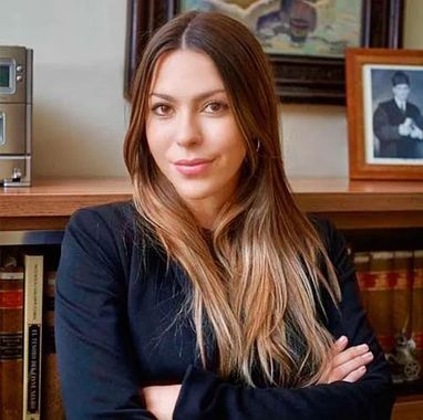 Mujer de pelo largo castaño, con los brazos cruzados, sentada en el interior de una casa frente a libros y fotos enmarcadas.